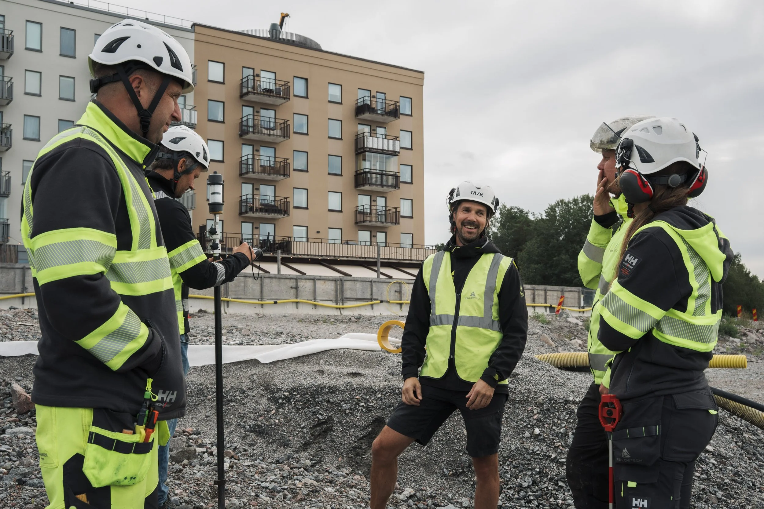 Construction workers in safety vests and helmets standing on an outdoor construction site, with a multi-story apartment building in the background.