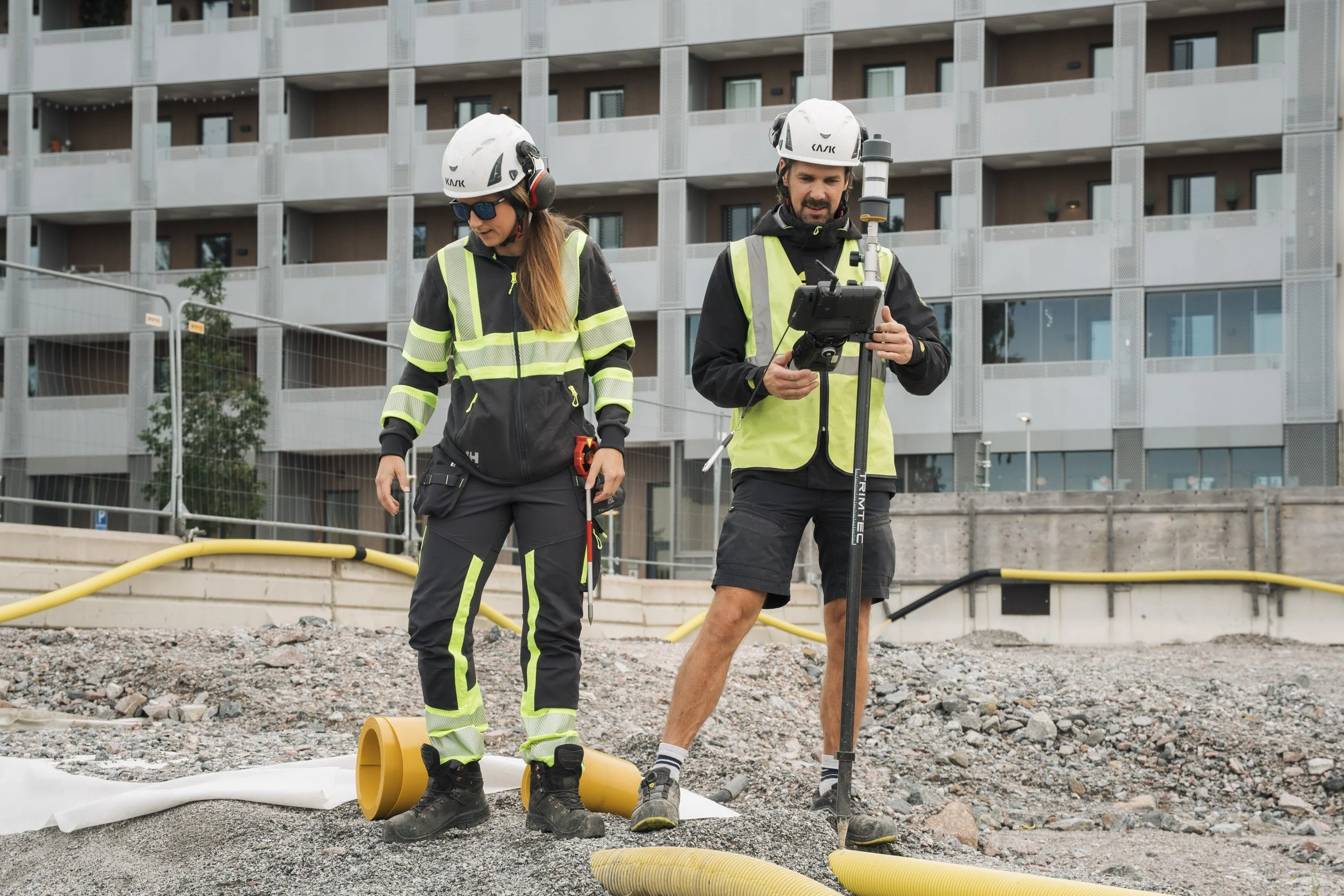 Two construction workers wearing safety helmets and reflective jackets working at a construction site with building apartment complex in the background. They are looking at equipment and inspecting the site, surrounded by construction materials and y