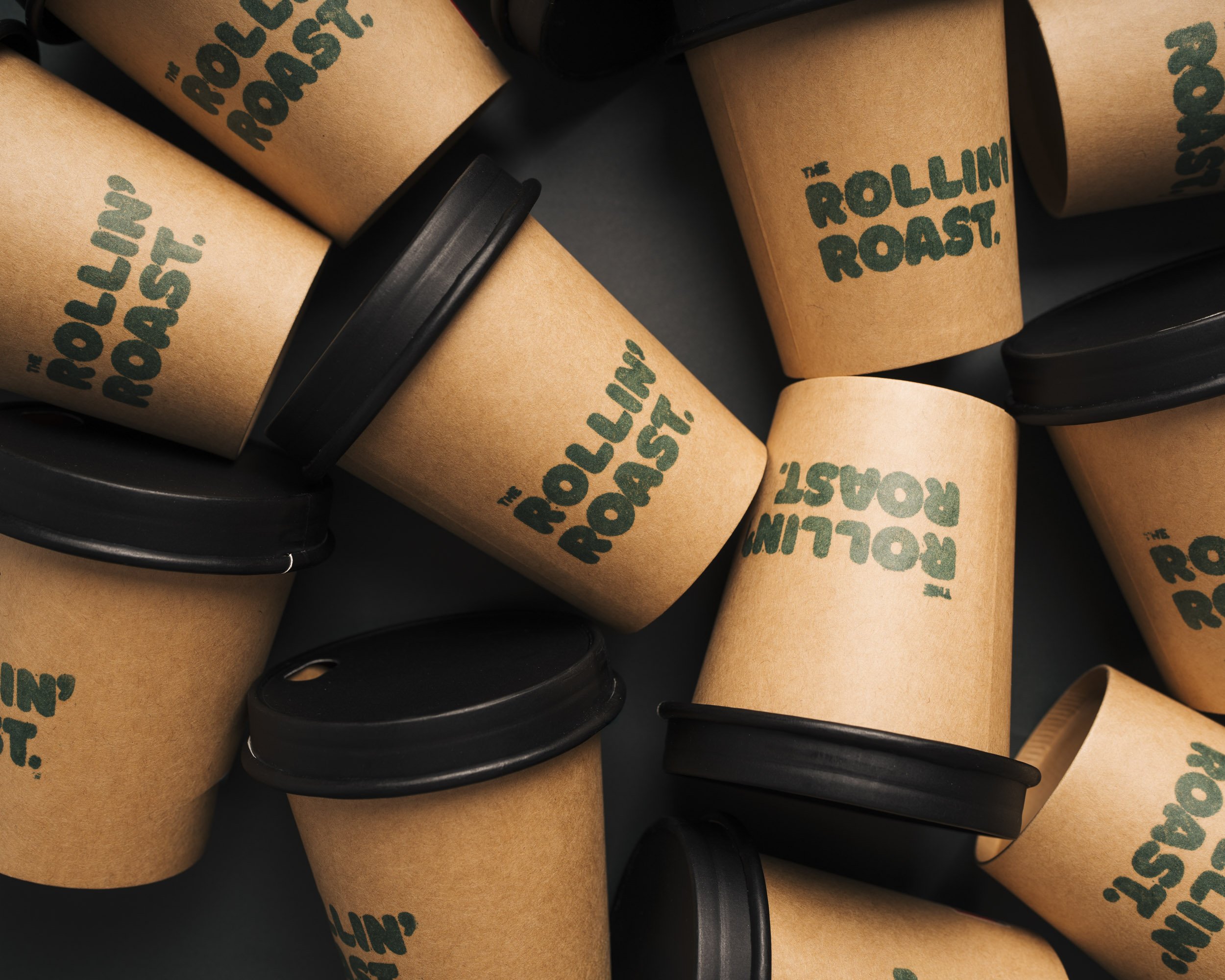Multiple reusable coffee cups with brown paper sleeves and black plastic lids, featuring the logo 'The Rollin' Roast' in green text.