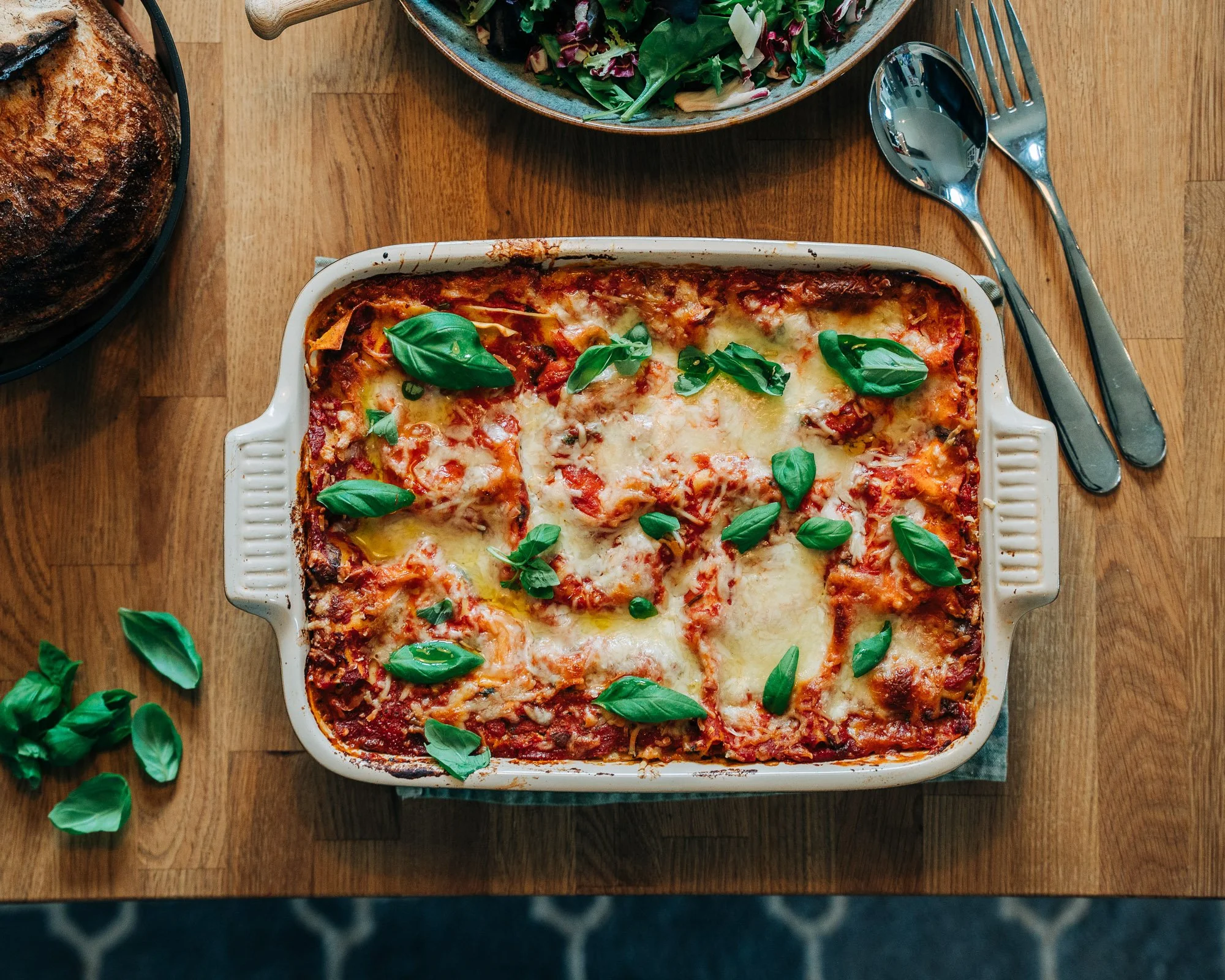 A casserole dish with baked lasagna topped with melted cheese and fresh basil leaves, on a wooden table with salad and utensils nearby.