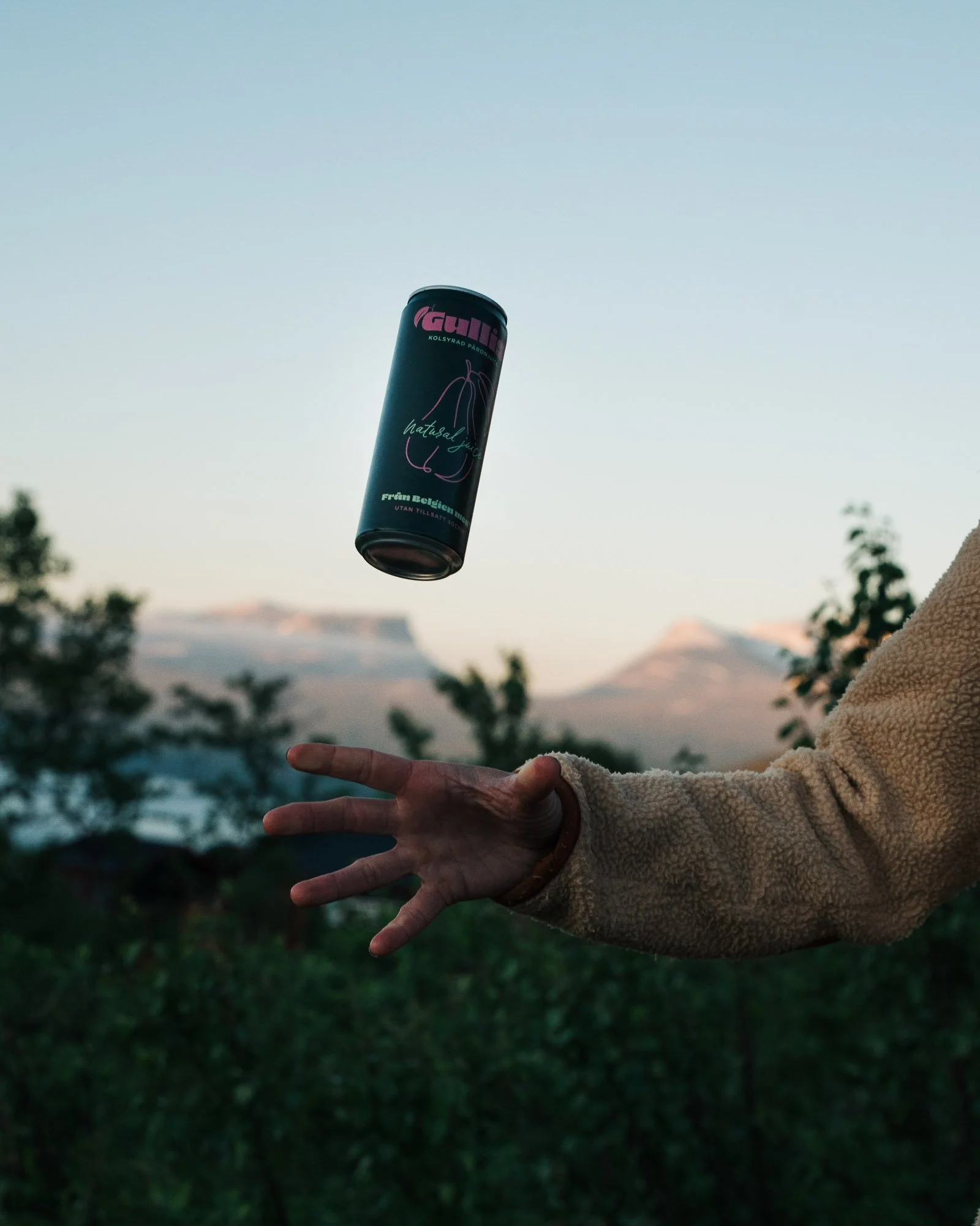 A person in a beige coat is tossing a can of GULD energy drink into the air outdoors during daytime with a blurred natural background.