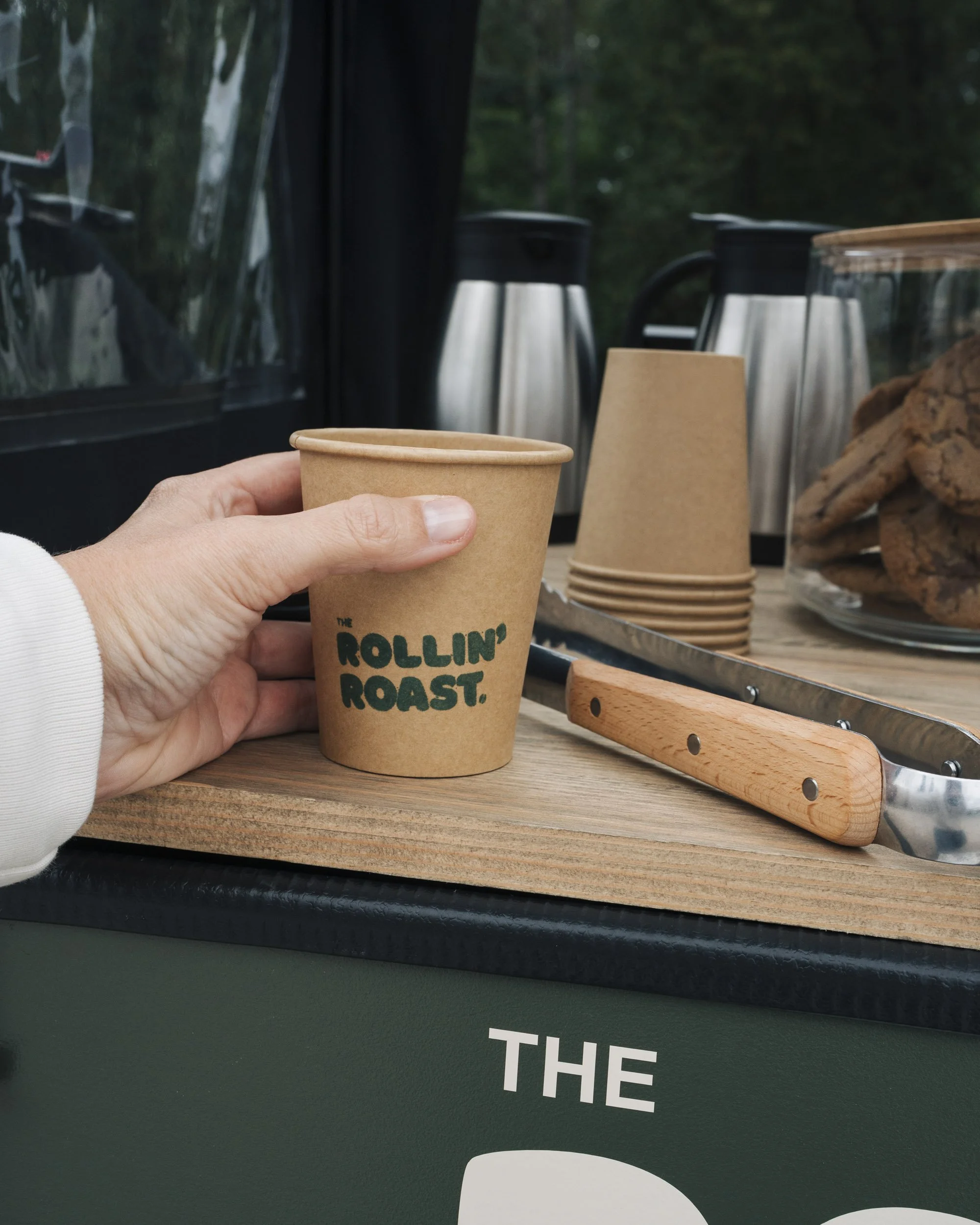 Hand holding a paper coffee cup with the phrase 'The Rollin' Roast' printed on it, set on a wooden surface with silver coffee urns, paper cups, cookies, and a glass jar of cookies in the background.