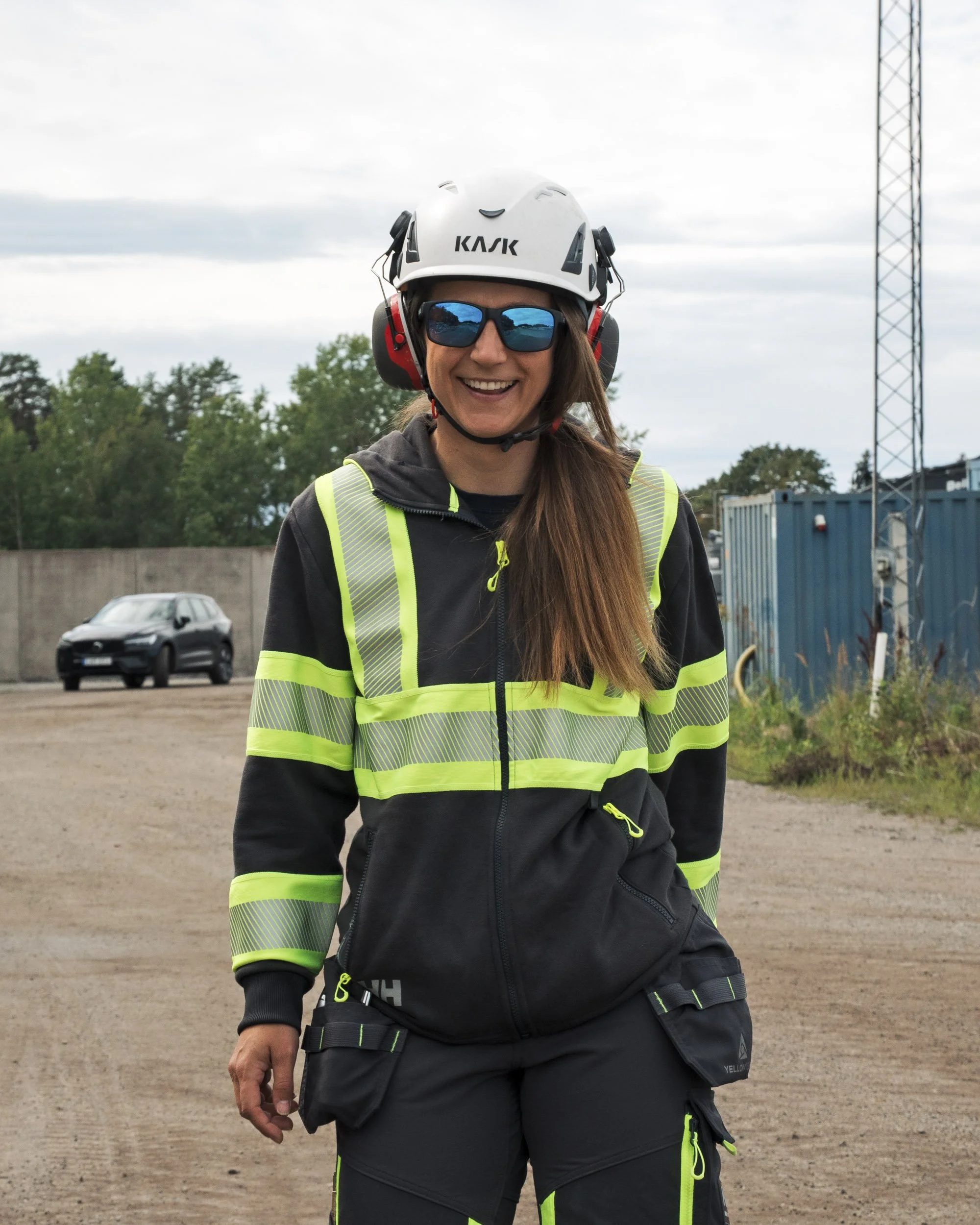 A smiling woman wearing a helmet, sunglasses, and a high-visibility jacket standing outdoors on a cloudy day. She has long brown hair and is dressed in black work pants with neon accents.