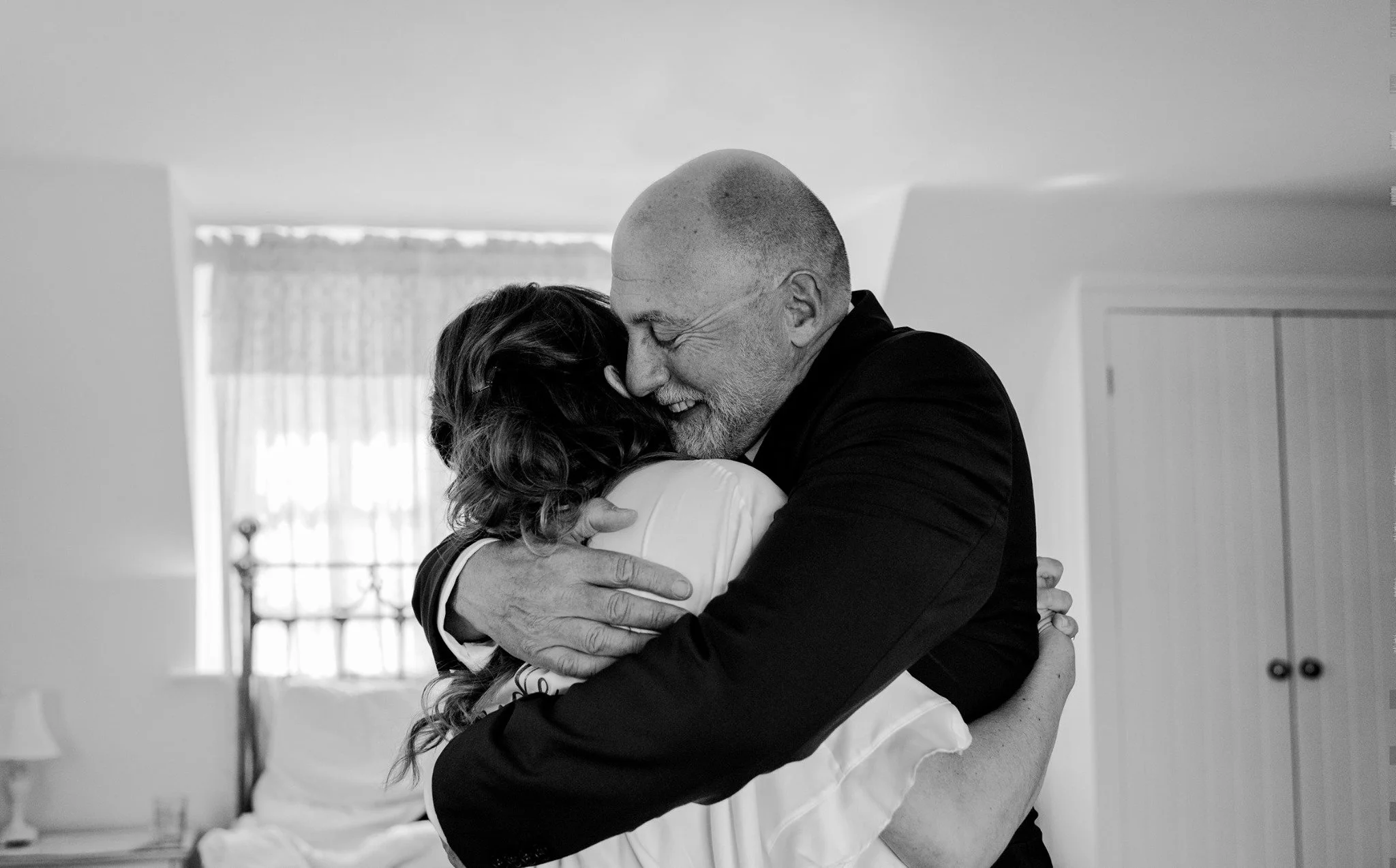 A man and woman hugging each other closely, smiling and with their eyes closed, in a bright room with a window in the background.