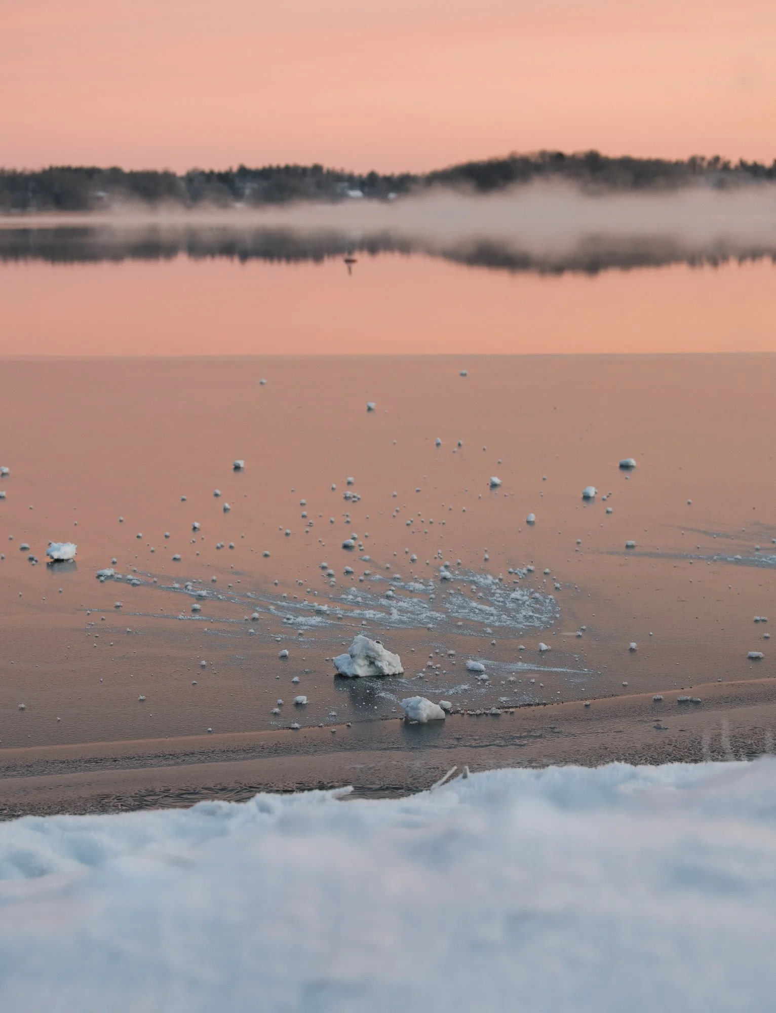 Frozen lake at sunset with small chunks of ice on the surface and mist rising from the water. Trees and hills in the distance under a pink sky.