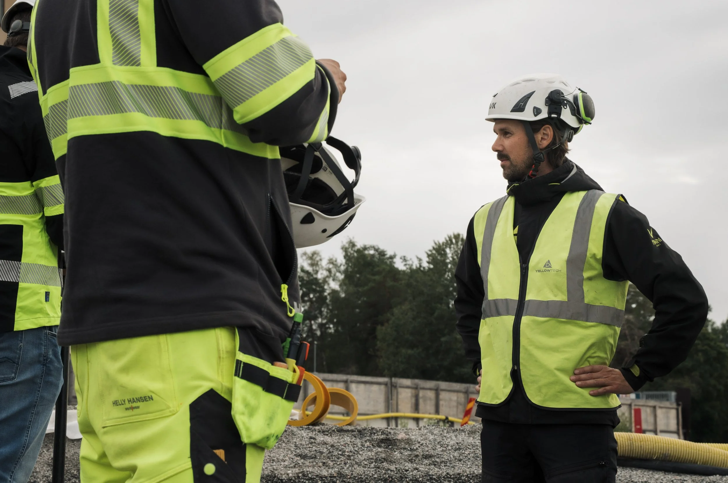 Group of construction workers wearing high-visibility yellow safety vests and helmets, discussing at a construction site with tools and equipment in the background.