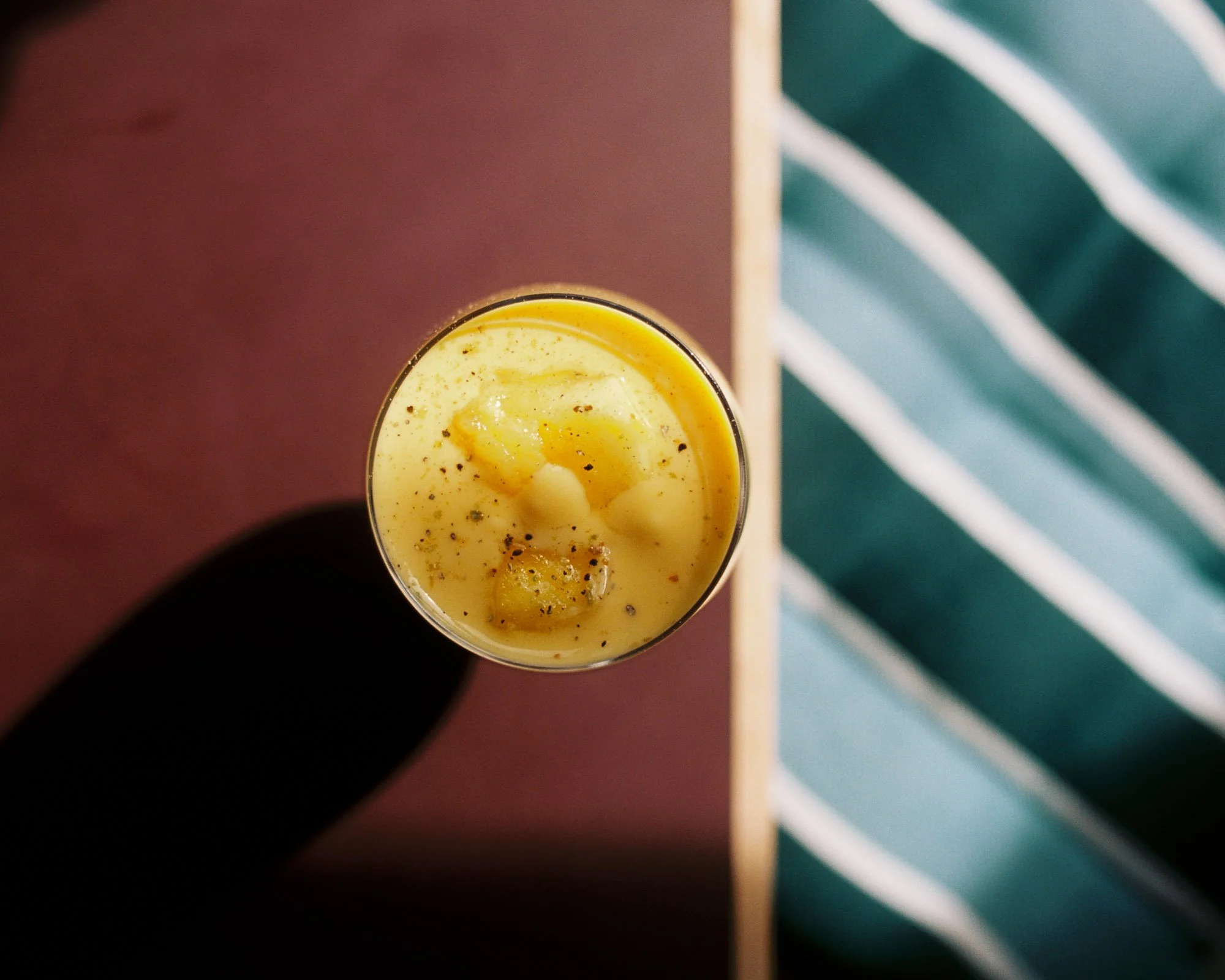 Top view of a glass filled with a creamy yellow dessert, garnished with black pepper and a small dollop of fruit or jam, placed on a wooden surface with a green and white striped cloth nearby.
