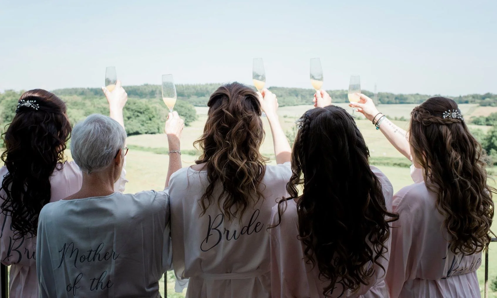 Group of women in matching robes celebrating with champagne outdoors, view of green fields and trees in the background.