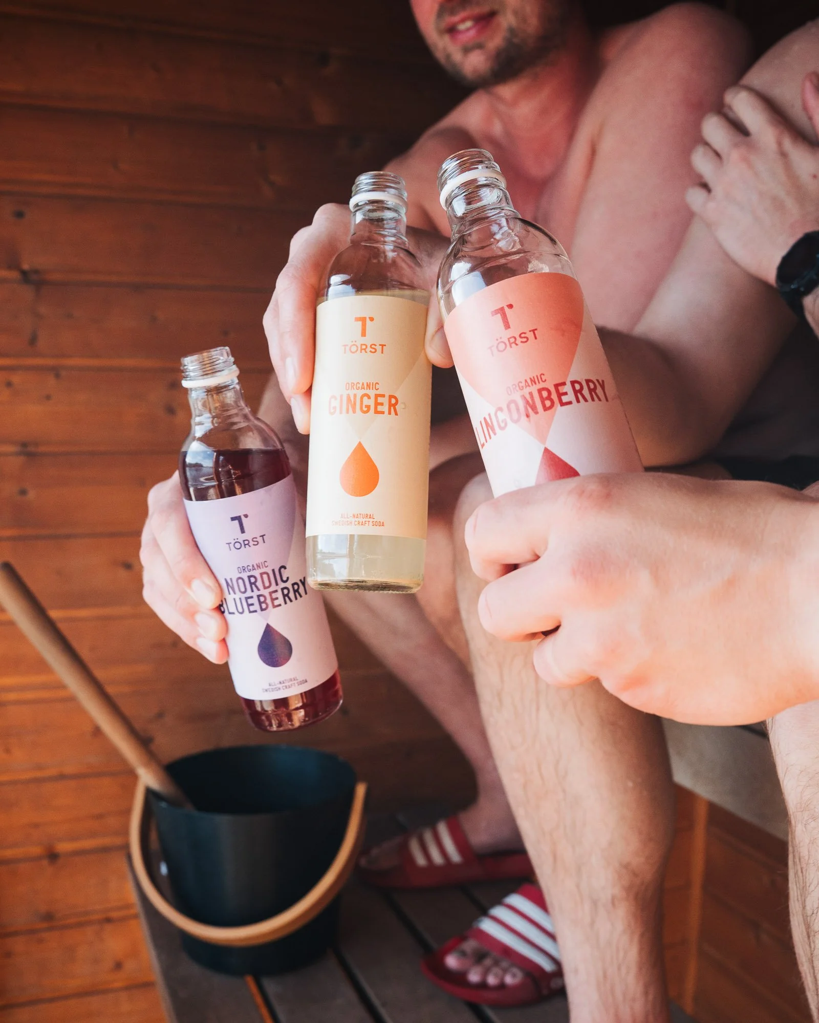 A shirtless man holding bottles of organic ginger and lingonberry soda, with a wooden wall background and a pair of red and white striped slides on his feet.