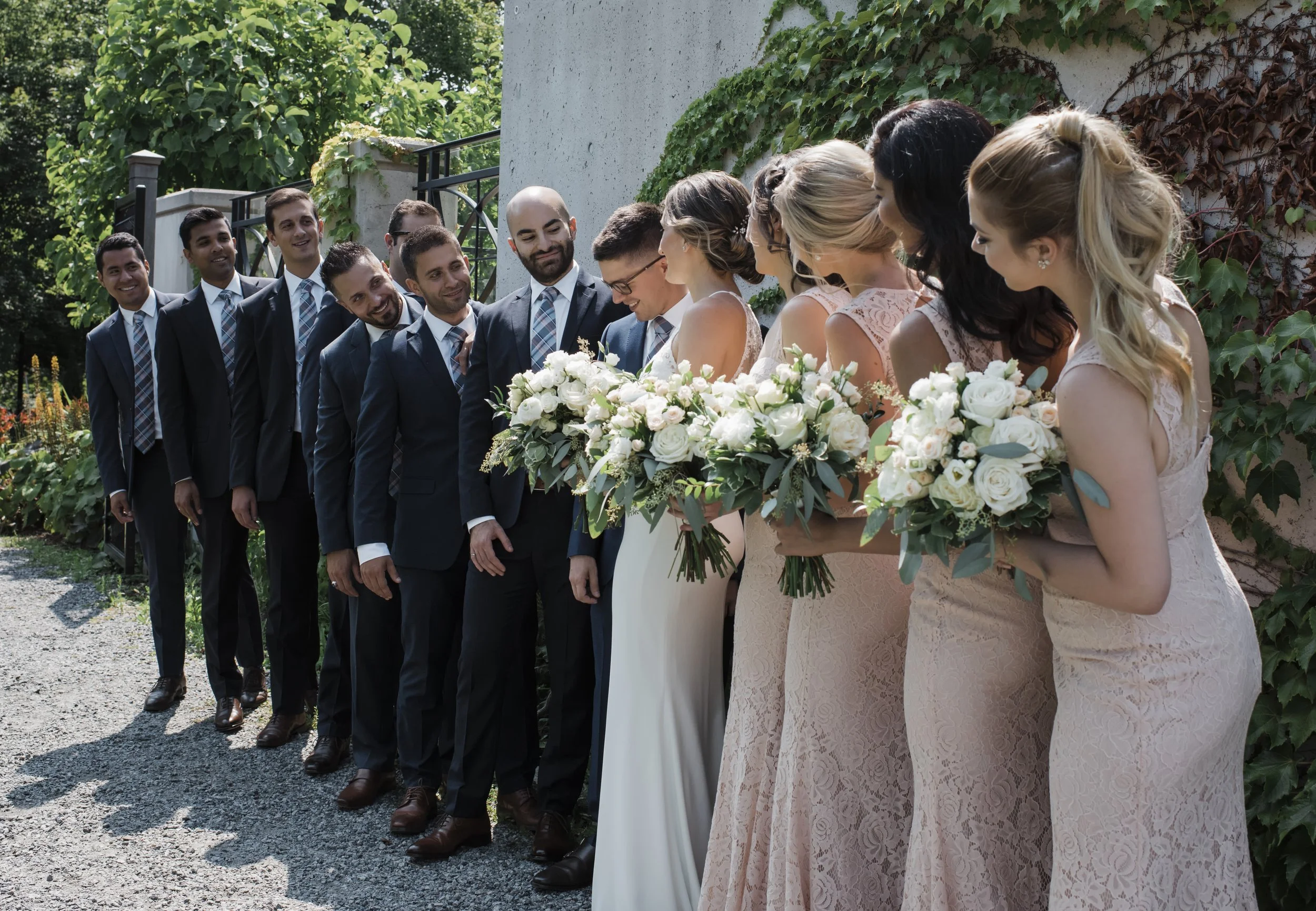A wedding party standing outdoors in a row, with the bride and bridesmaids in front holding bouquets of white flowers, and the groom and groomsmen behind wearing suits and ties, during daylight.