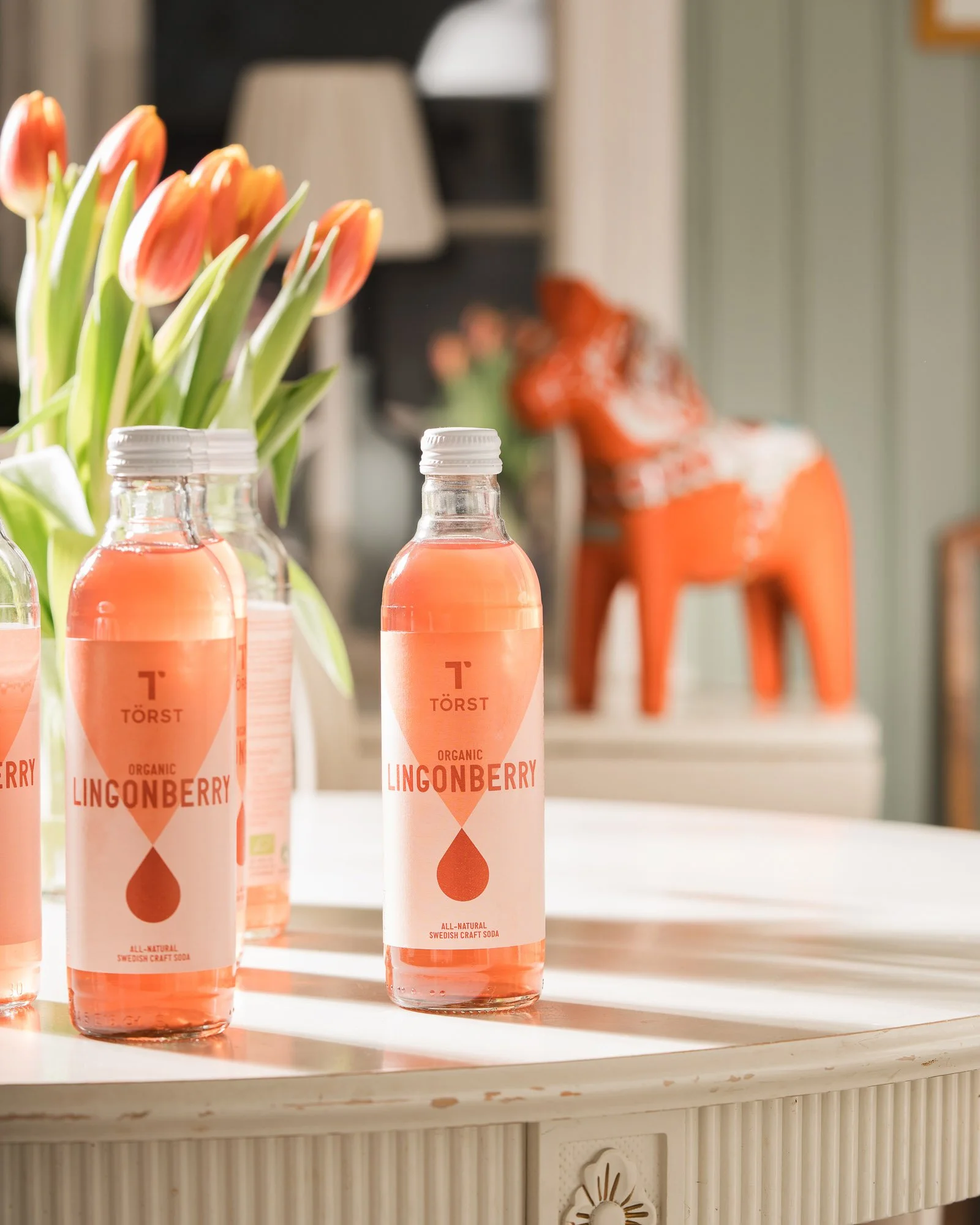 Four bottles of Lingonberry Swedish craft soda Törst  on a white table, with a bouquet of orange tulips in the background.