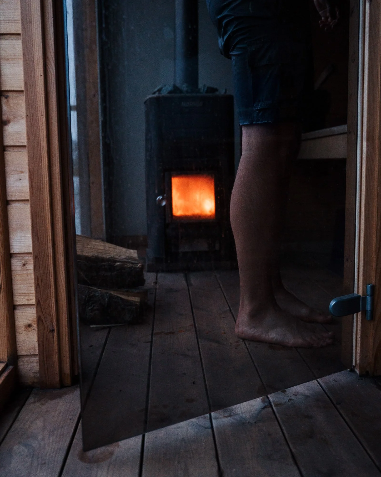 Person standing in sauna with wood stove glowing orange, wooden interior, and tiled floor.