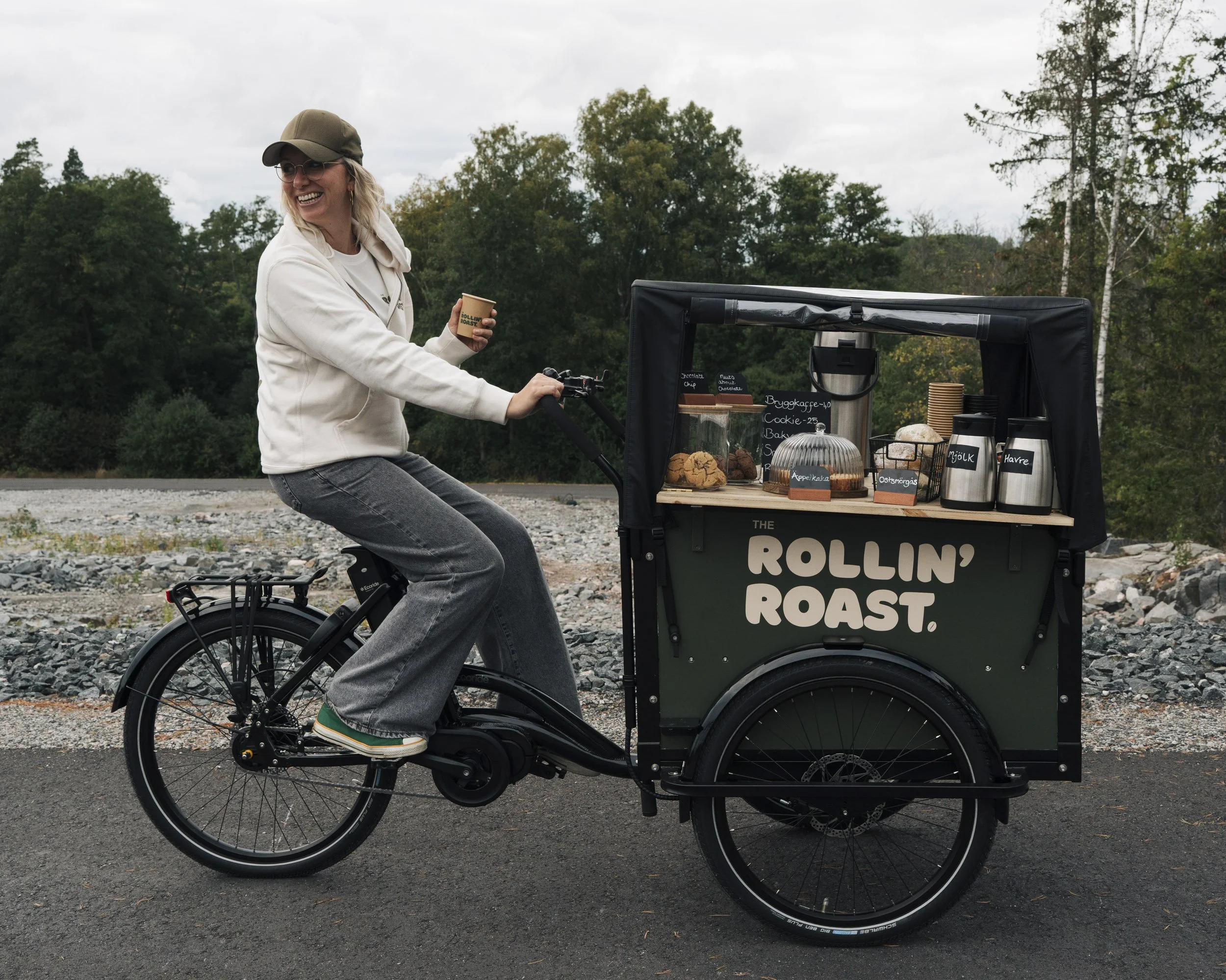 A woman wearing a cap, glasses, and a white jacket rides a black cargo bicycle with a snack cart attached, outdoors in a natural setting with trees and cloudy sky.