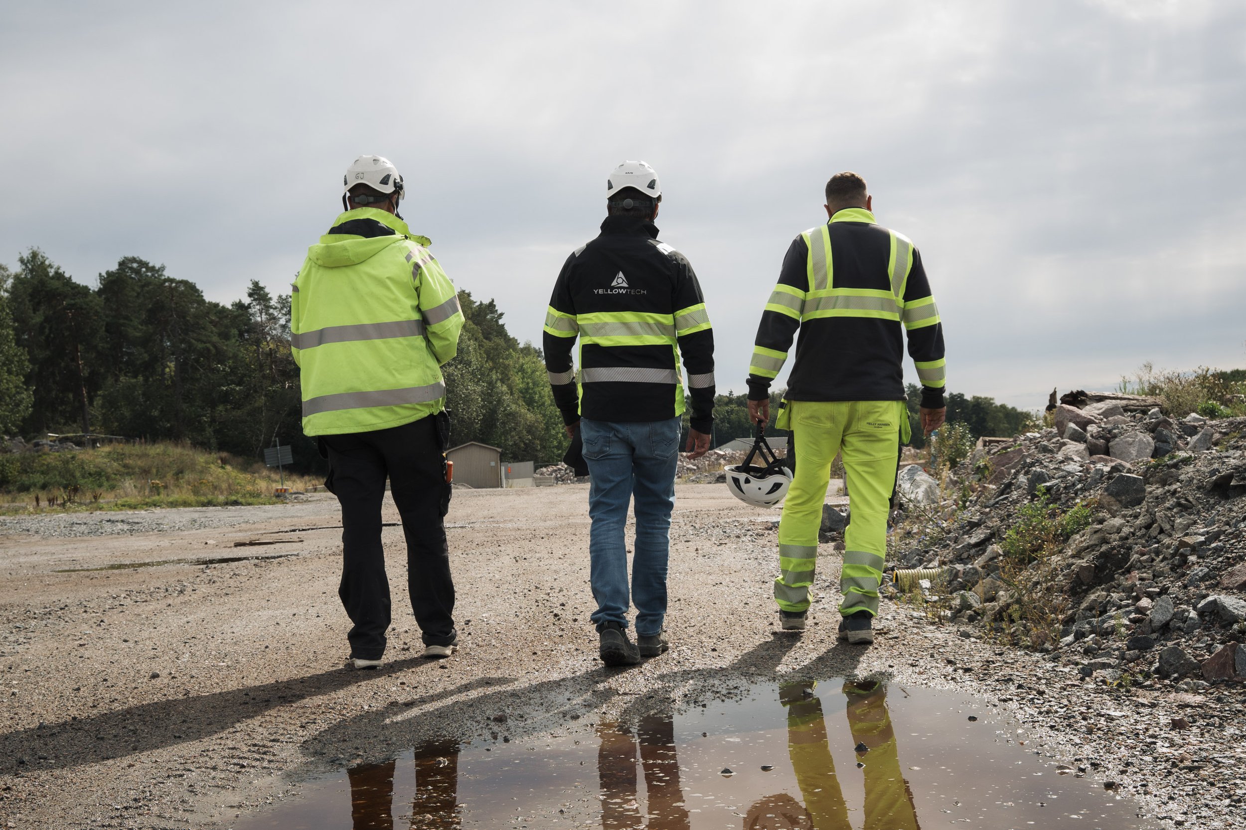Three construction workers wearing safety vests and helmets walk on a gravel road near a construction site with piles of rocks and a puddle reflecting their images.