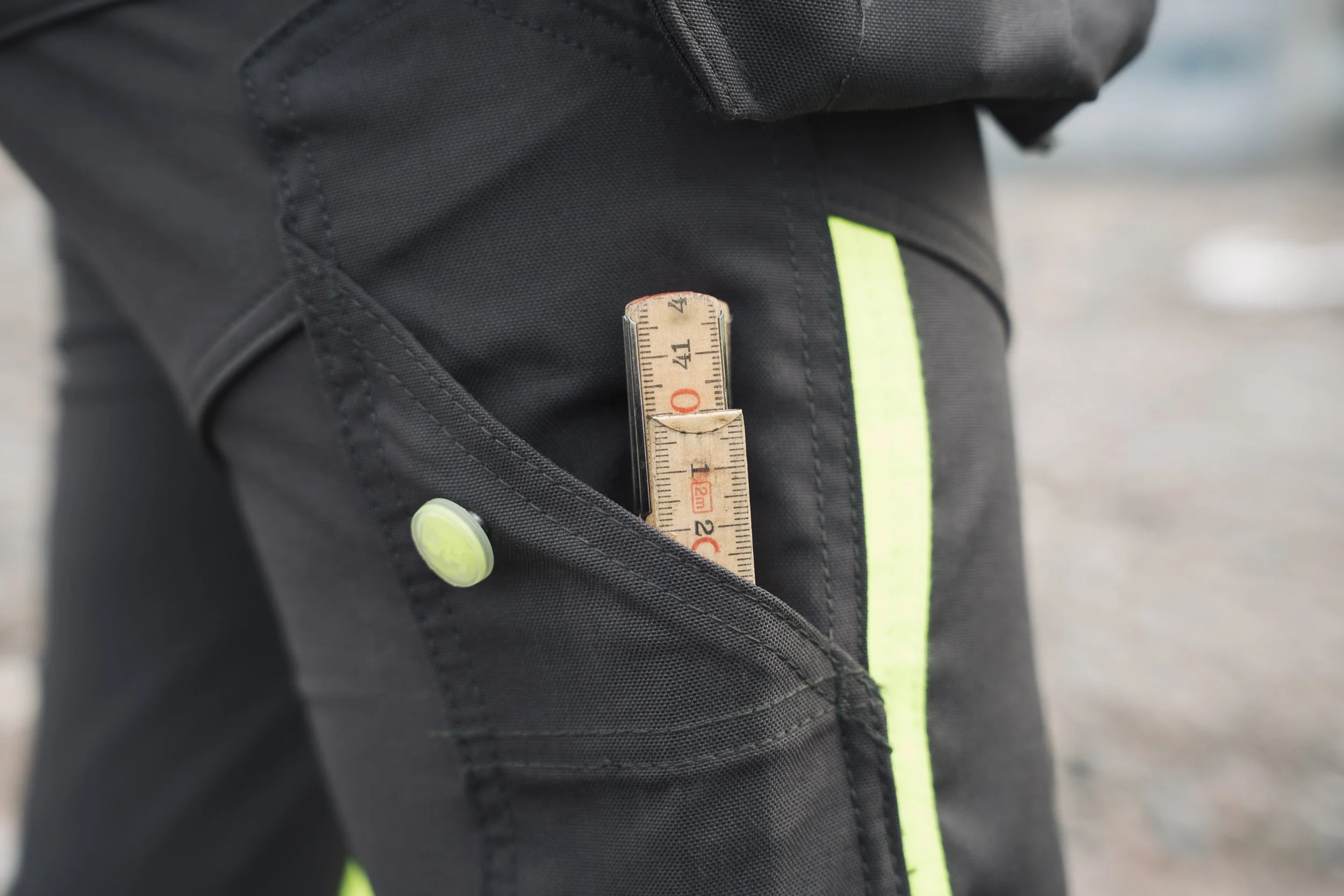 Close-up of a black backpack with a yellow-green stripe, featuring a measuring tape tucked into one of its compartments.