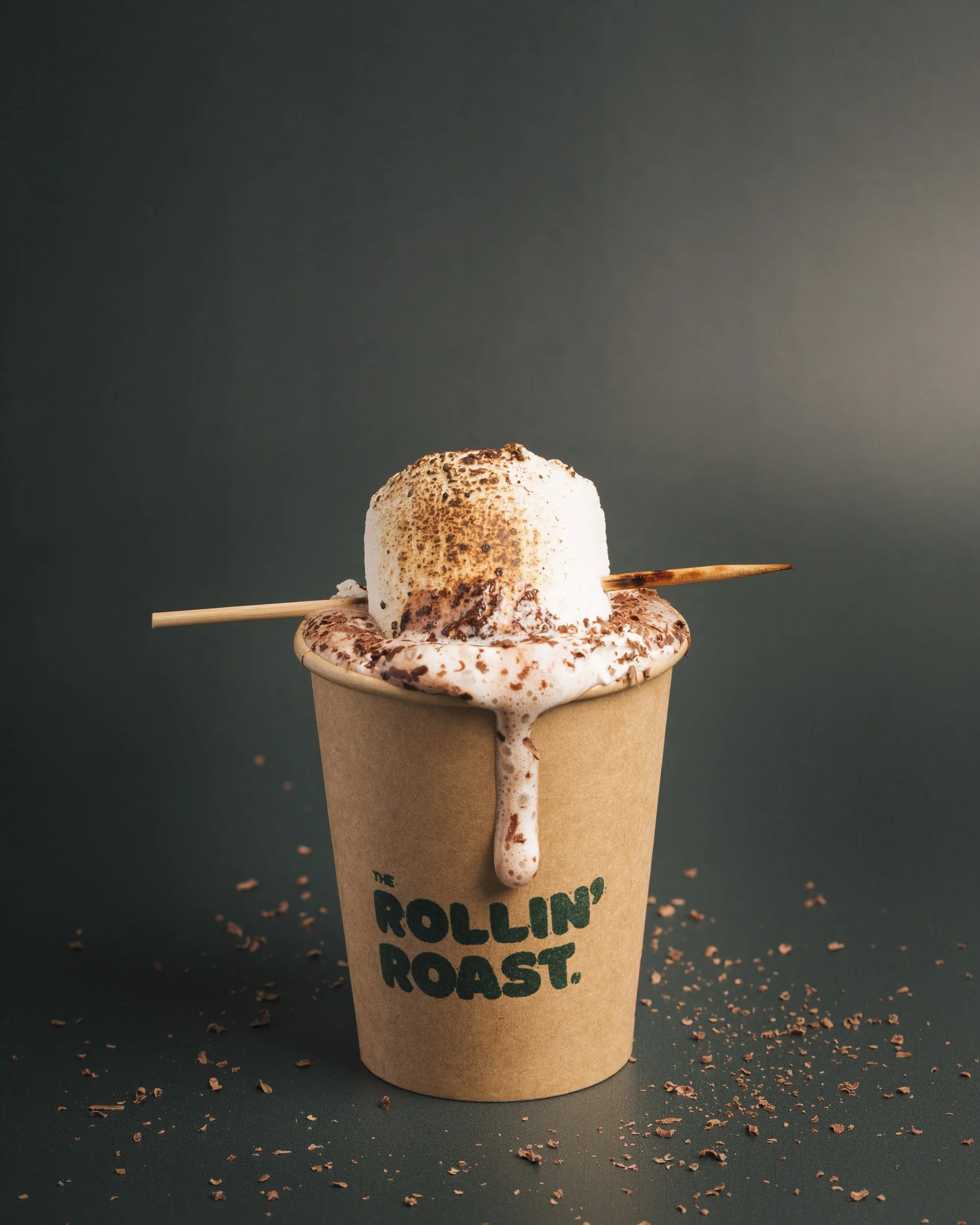A paper coffee cup with a logo that reads 'The Rollin' Dust'. The cup is filled with a dessert topped with whipped cream and chocolate shavings, with a spoon appended through the whipped cream. The dessert appears to be a marshmallow or similar treat