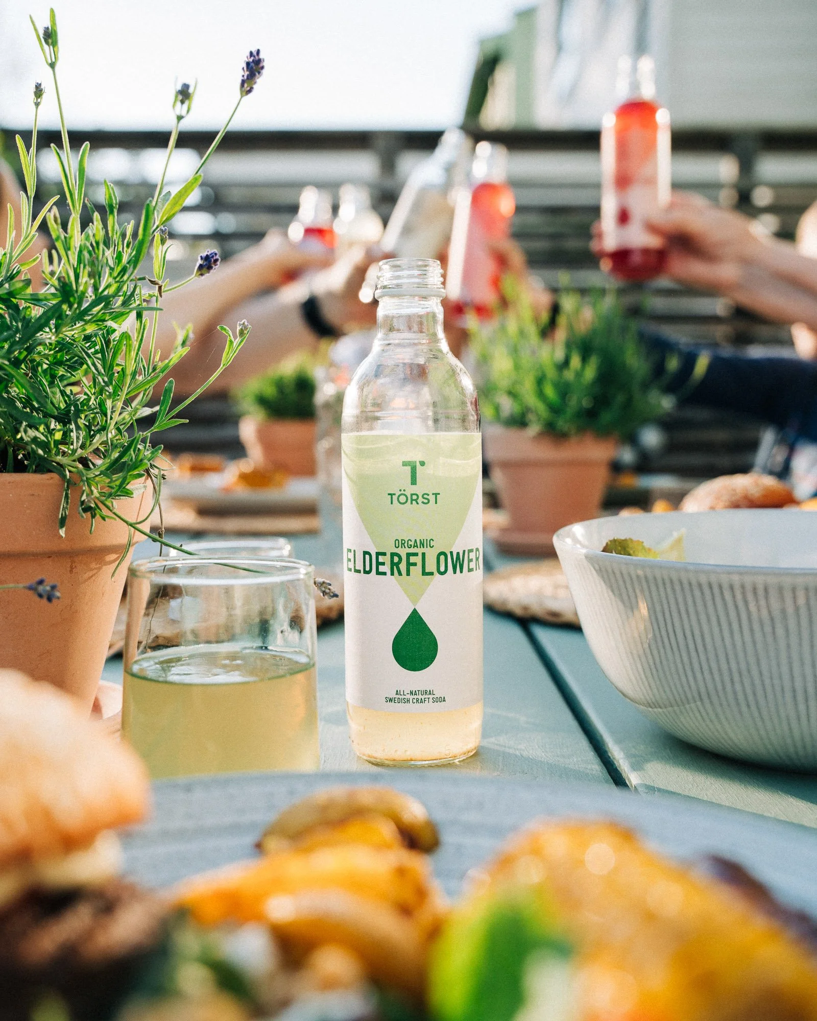 A table with potted lavender plants, glasses of water, and a drink bottle of Törst Elderflower amidst food items, with a blurred background of people raising bottles during a gathering.