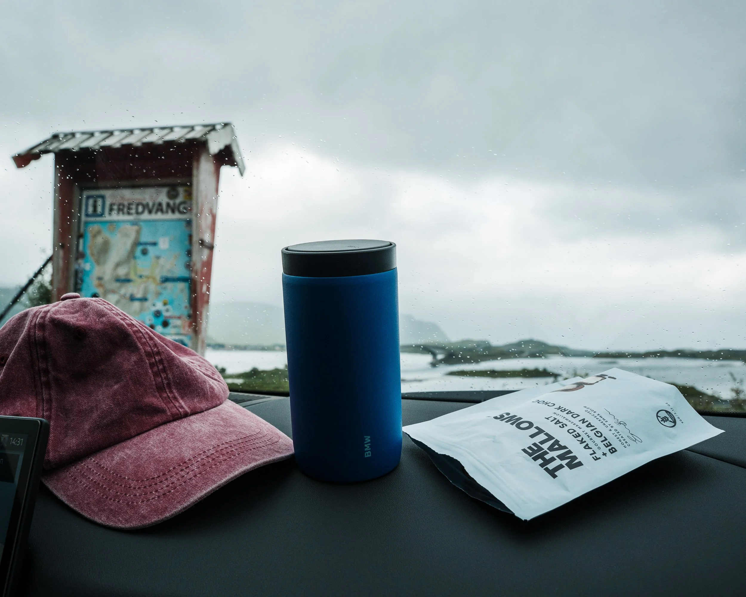 Inside a vehicle, view of a pink cap, a blue water bottle, a paper bag, a red and black sign boarding a bridge, and a stormy cloudy sky through the windshield.