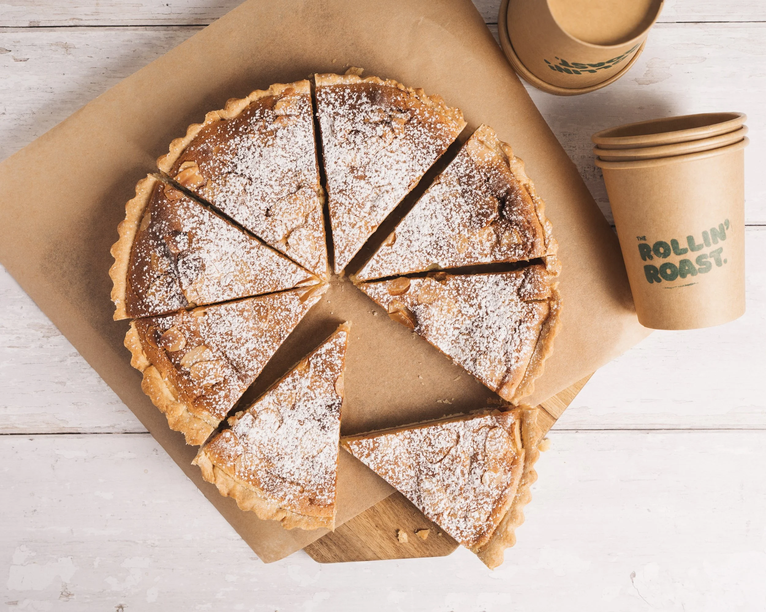 A partially sliced caramel tart dusted with powdered sugar on brown parchment paper, with a stack of brown coffee cups and a to-go coffee cup labeled 'The Rolling Roast' on a white wooden surface.