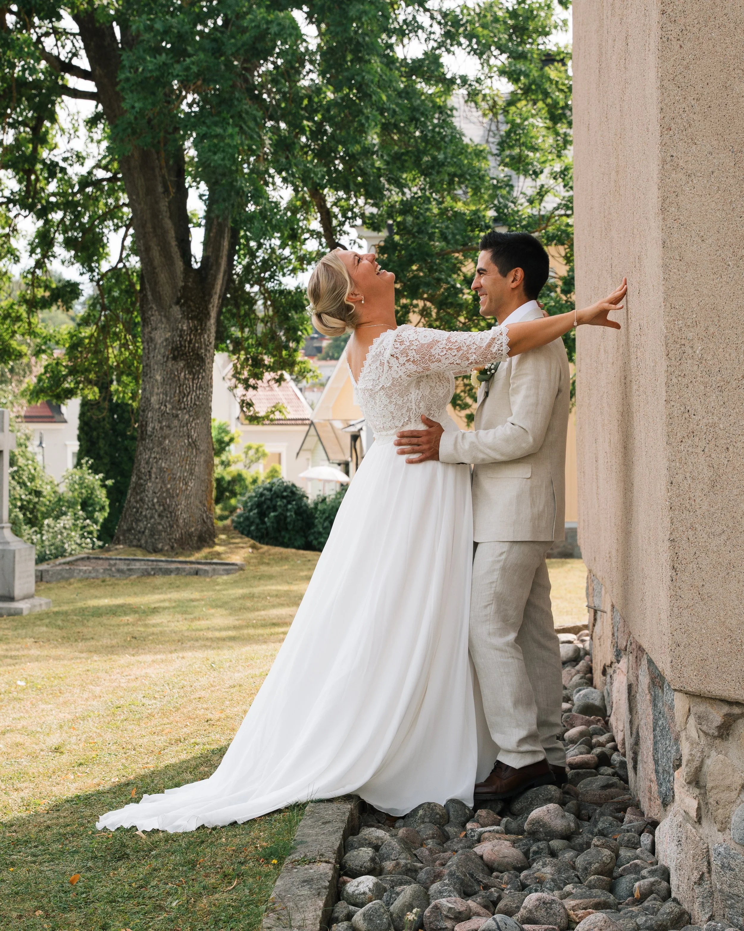 A bride and groom laughing and kissing outdoors near a textured beige wall with rocks at the base. They are dressed in wedding attire, with the bride in a white lace dress and the groom in a light-colored suit. Tall trees and a residential neighborho