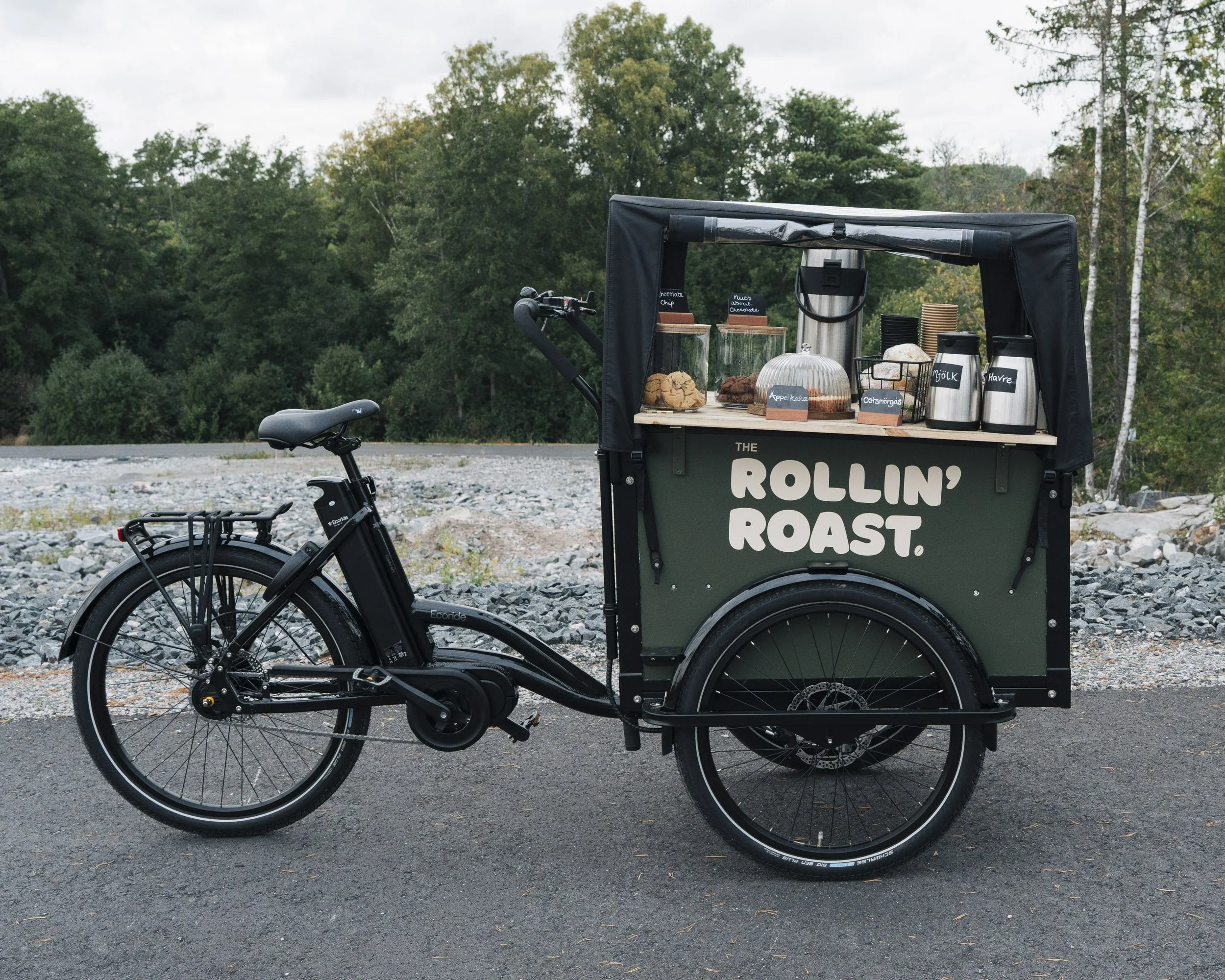 A bicycle with a food cart attached to it, which has baked goods and drinks on display, parked on a gravelly outdoor area with trees in the background.