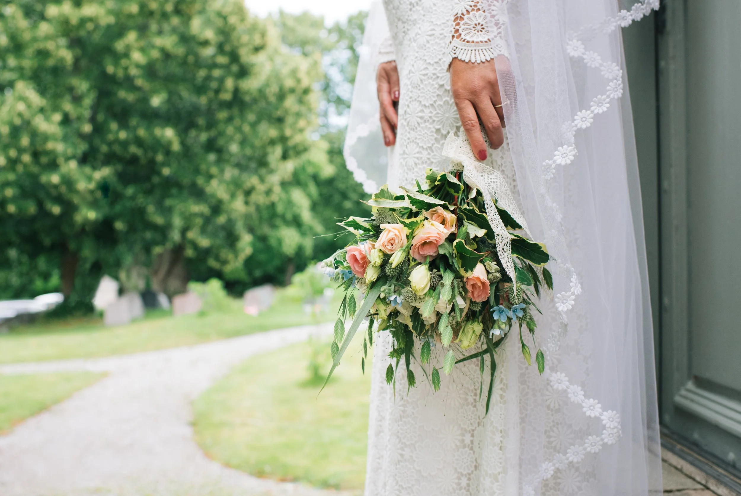 Close-up of a bride holding a bouquet of light pink roses and greenery outdoors near a house with a gray wall and white lace dress with floral embroidery.