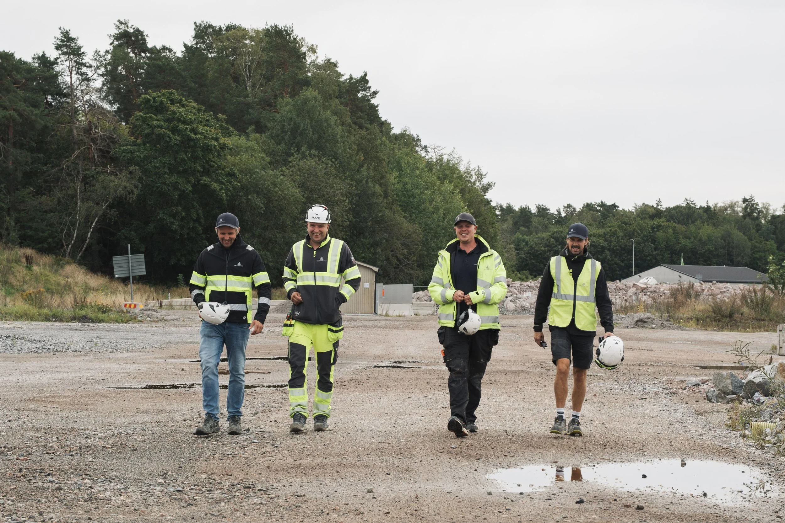 Four men walking on a gravel path in a construction or outdoor work site, wearing safety gear including helmets and high-visibility jackets, with a backdrop of trees and construction equipment.