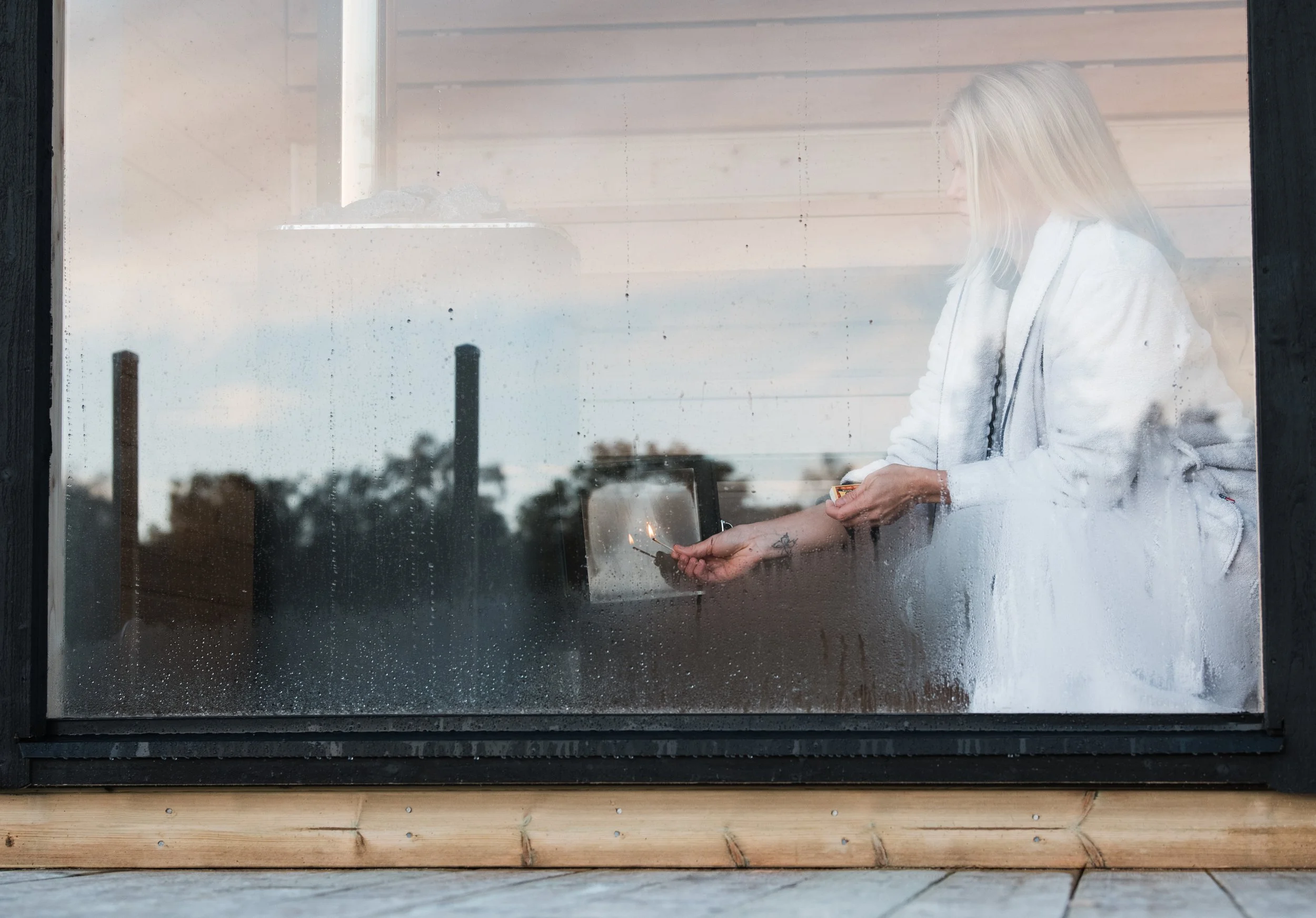 A person with long blonde hair, wearing a white hoodie, is inside looking out a rain-covered window and holding a matchstick.
