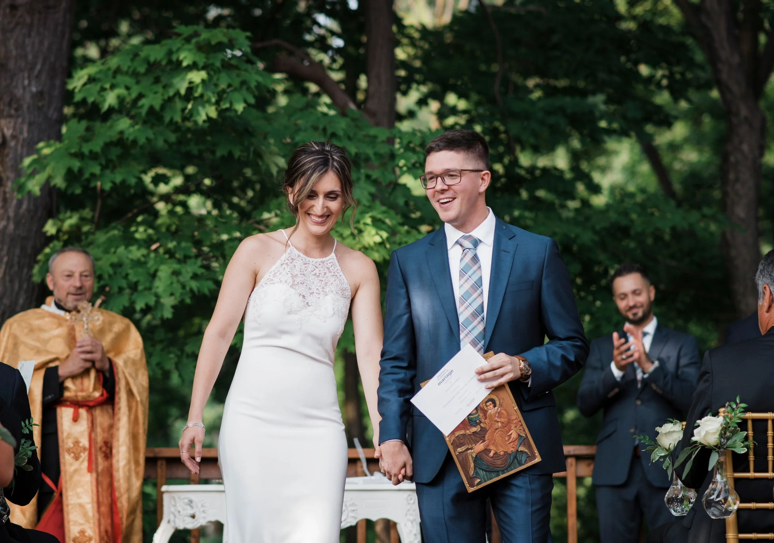 A bride and groom exchanging vows at an outdoor wedding ceremony, with family and friends present, under a canopy of green trees.