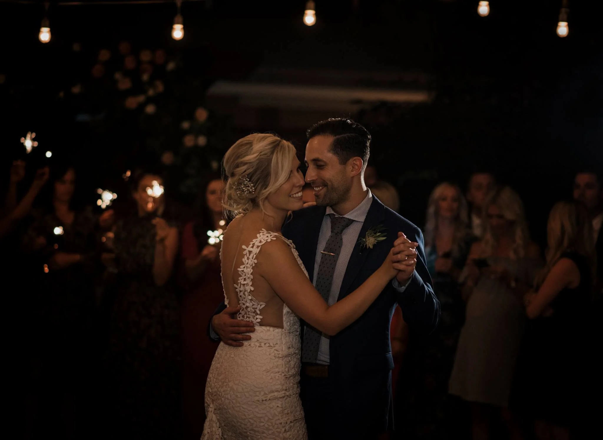A bride and groom share a dance at their wedding reception, surrounded by guests, with warm hanging lights creating a cozy atmosphere.