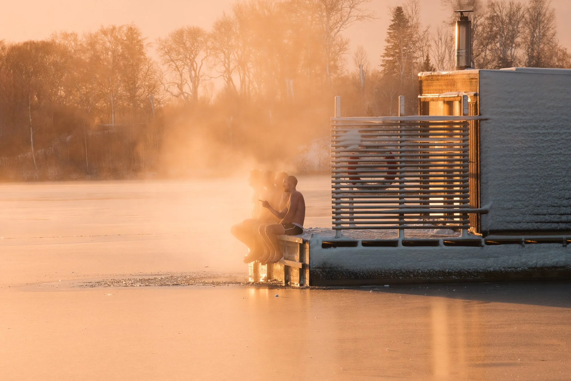 People sitting on a dock by a frozen lake during a sunset or sunrise, with steam rising from the water, and trees in the background.