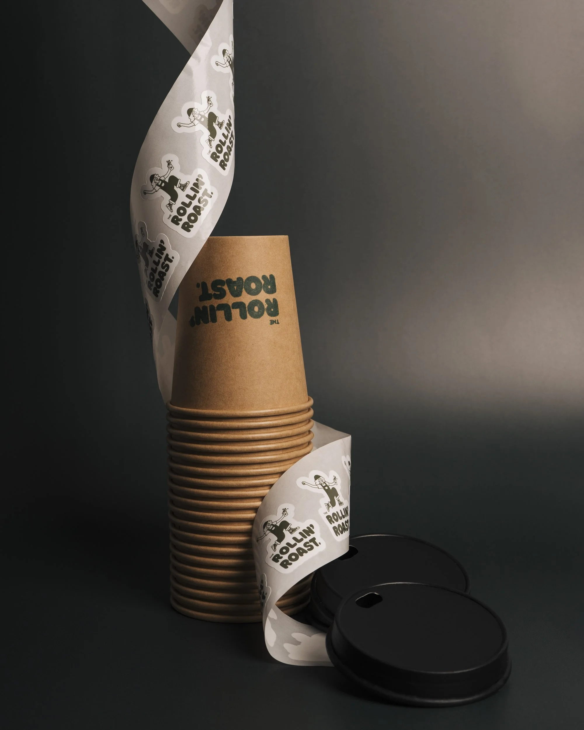 Stacked brown paper coffee cups with black plastic lids, some cups have green text that says 'The Rolling Roast,' and there are paper wrappers with cartoon illustrations and the text 'The Rolling Roast' in black on a white background, against a dark 