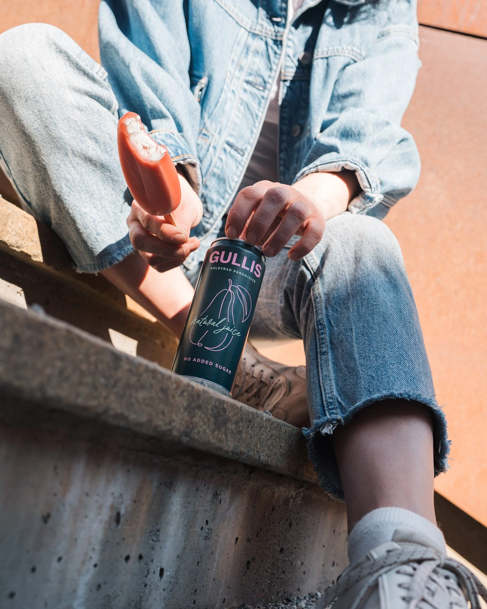 A person sitting on a concrete ledge wearing light blue denim jeans, a denim jacket, and white sneakers. They are holding a can of Gullis brand natural juice with a chocolate popsicle in their other hand.