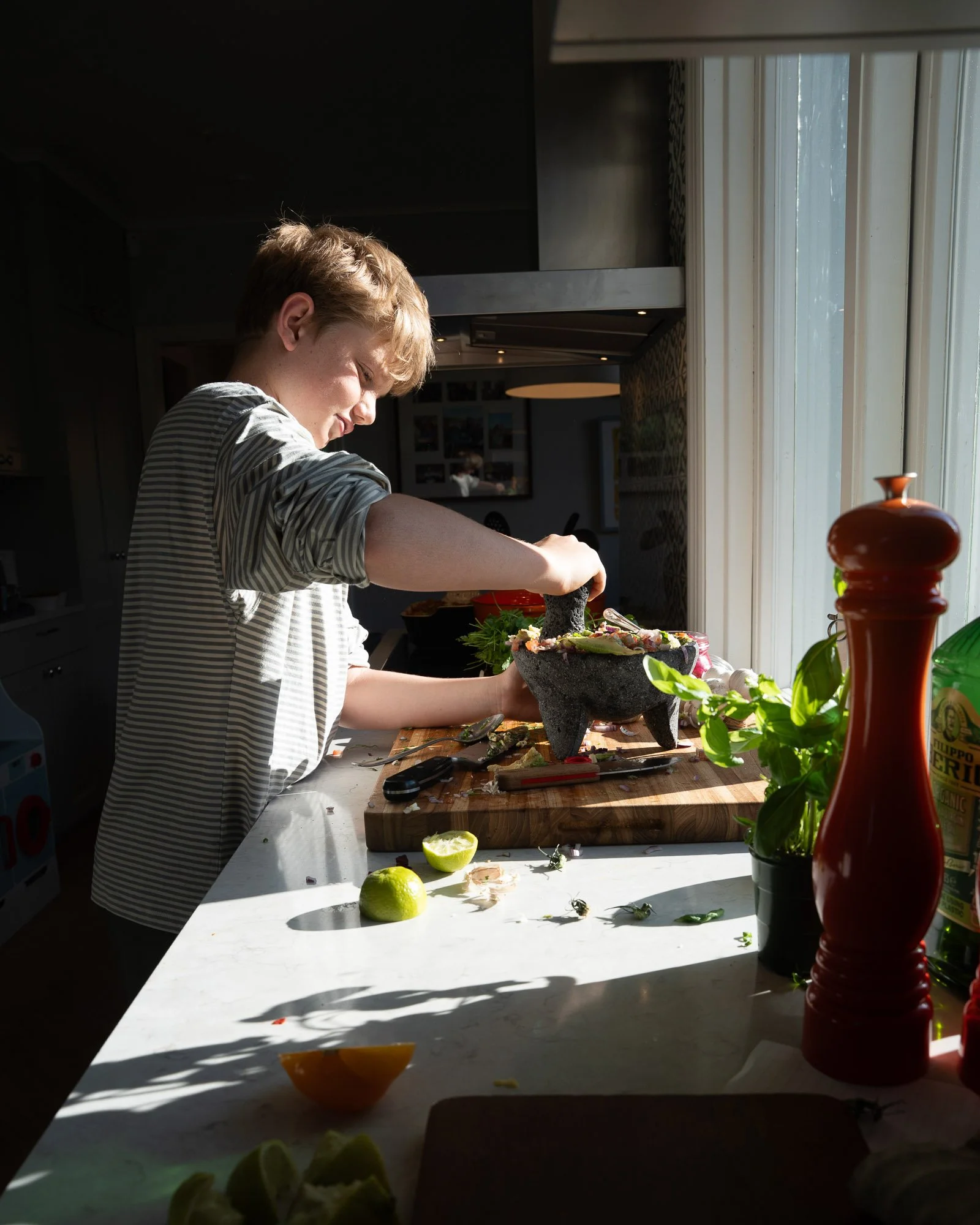 A boy preparing food in a kitchen by chopping vegetables on a wooden cutting board, with sunlight streaming through a window.