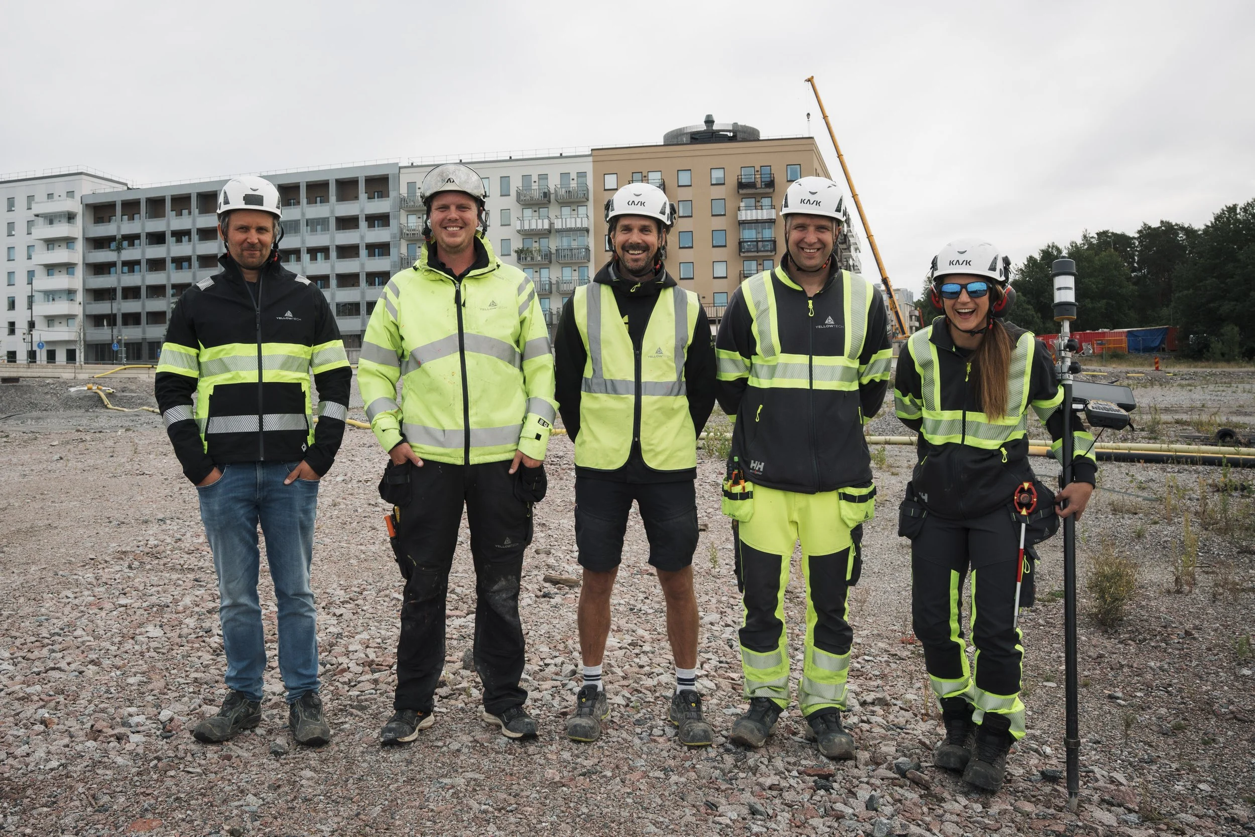 Group of five construction workers standing on a gravel site in front of modern apartment buildings, wearing safety helmets and high-visibility jackets, smiling and posing for the photo.