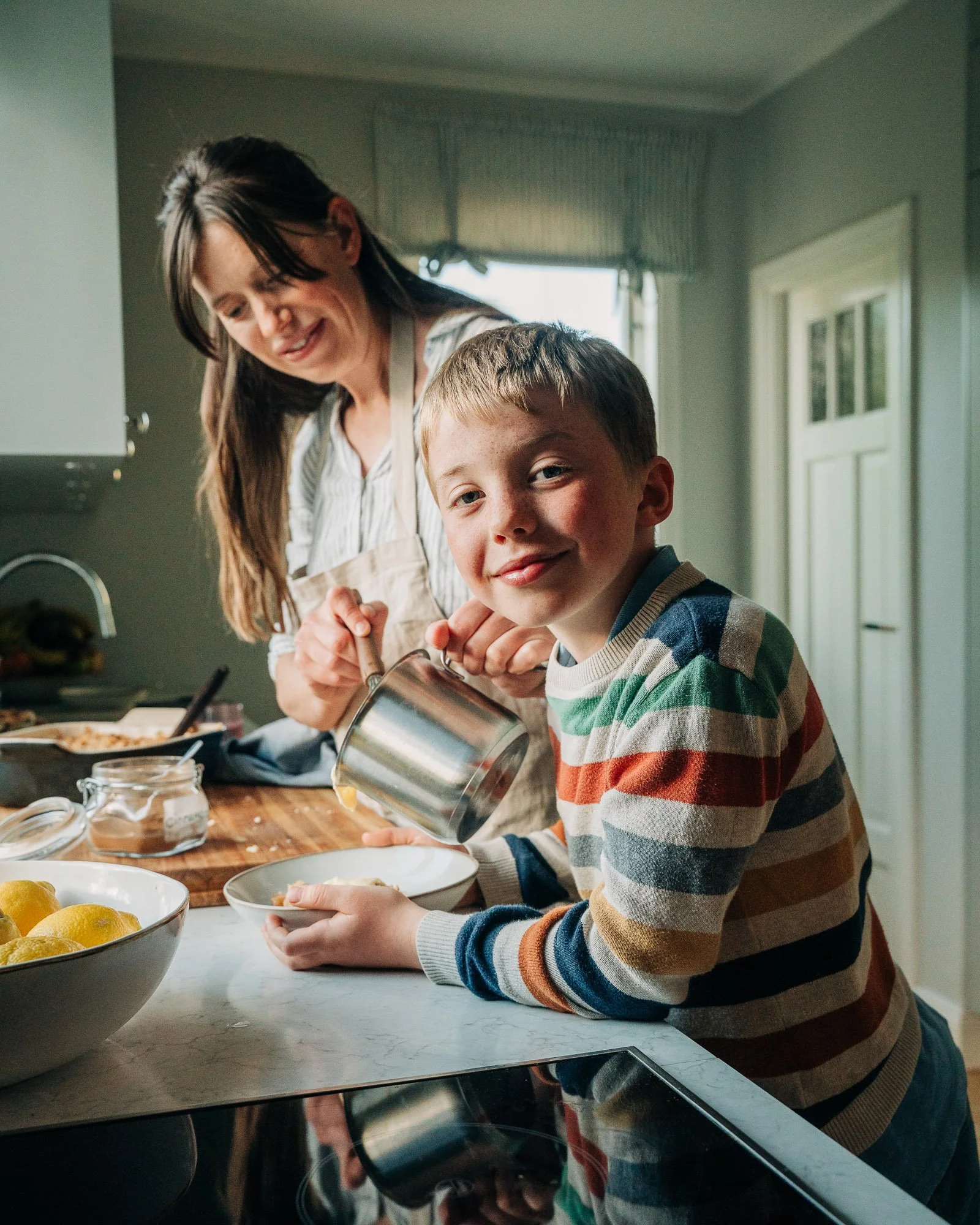 A woman and a young boy preparing food in a kitchen. The boy is pouring ingredients into a bowl, and the woman is smiling and looking down. The kitchen has a window with a curtain and framed pictures on the wall. On the counter are bowls of lemons an