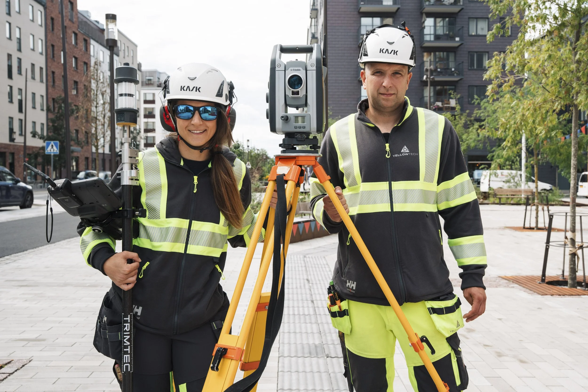 Two surveyors in safety gear standing outdoors with surveying equipment, urban background with buildings and trees.