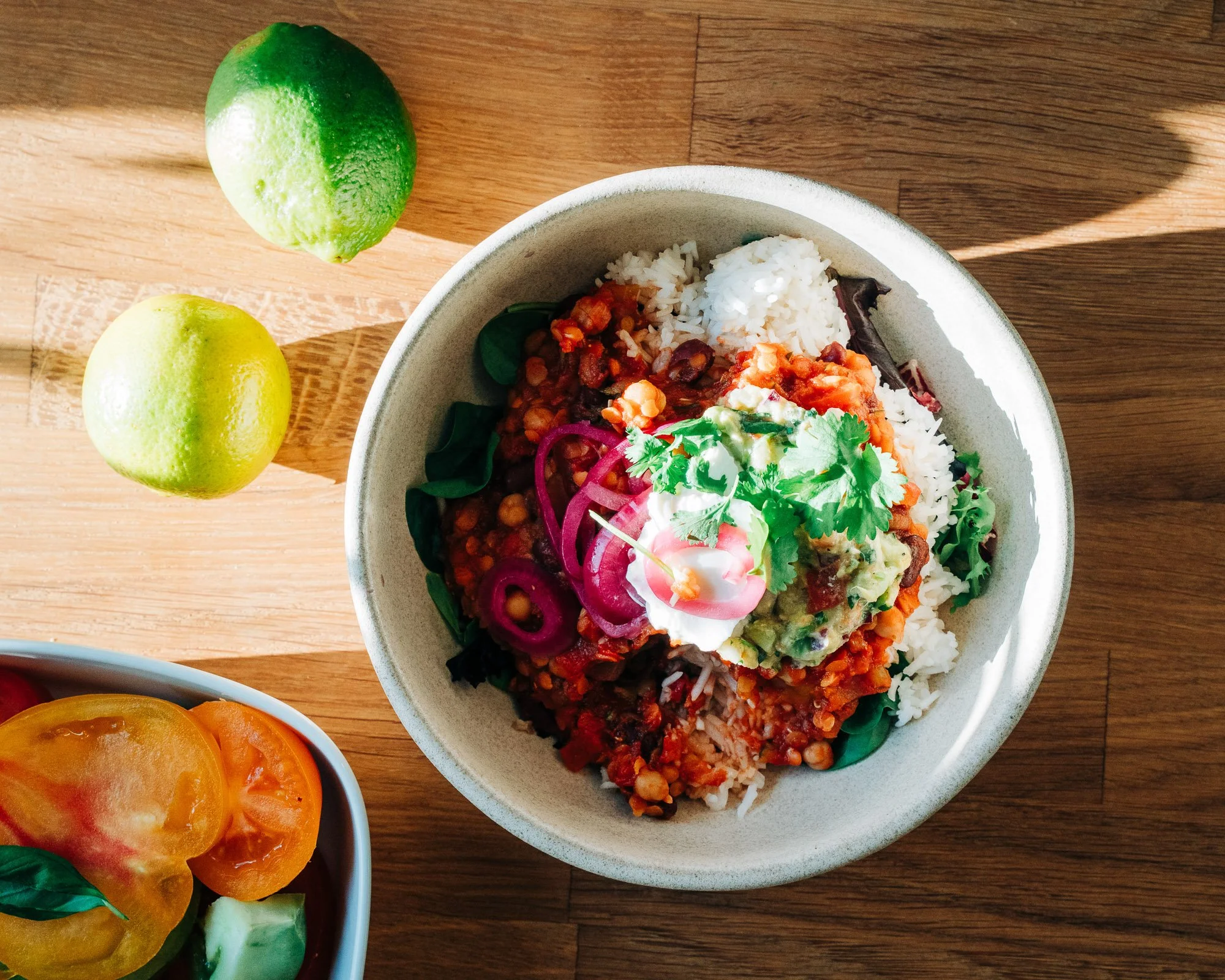 A bowl of rice topped with chili, salsa, and chopped cilantro, with sliced tomatoes on the side and two limes on the wooden table.