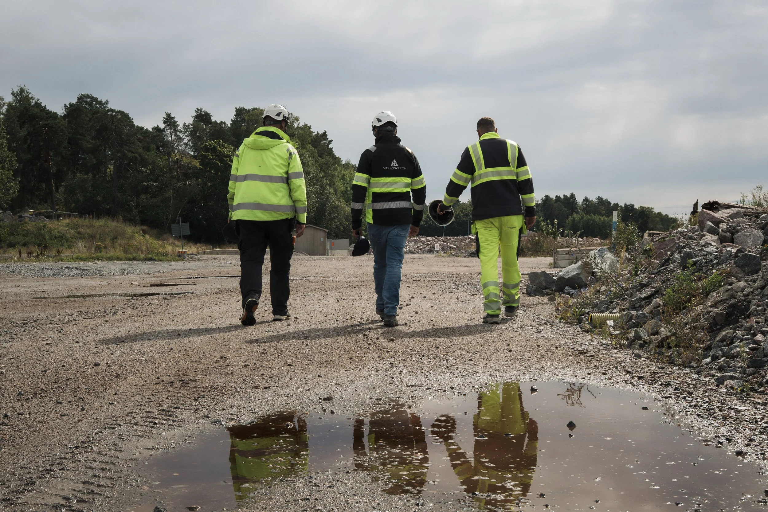 Three men in safety vests and helmets walking on a dirt pathway beside a pile of rocks and a puddle, with trees in the background and cloudy sky overhead.
