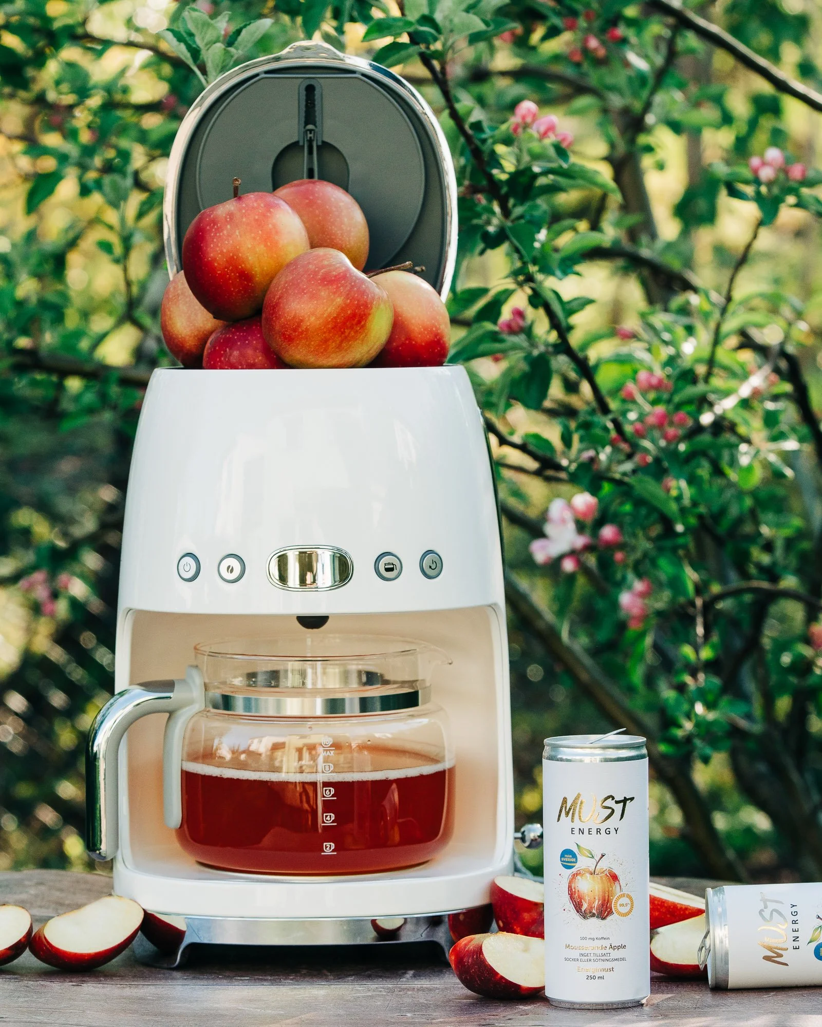 A cold brew coffee maker with fresh apples on top, outdoors surrounded by green foliage and pink flowers, with apple-flavored energy drinks and apple slices around it.