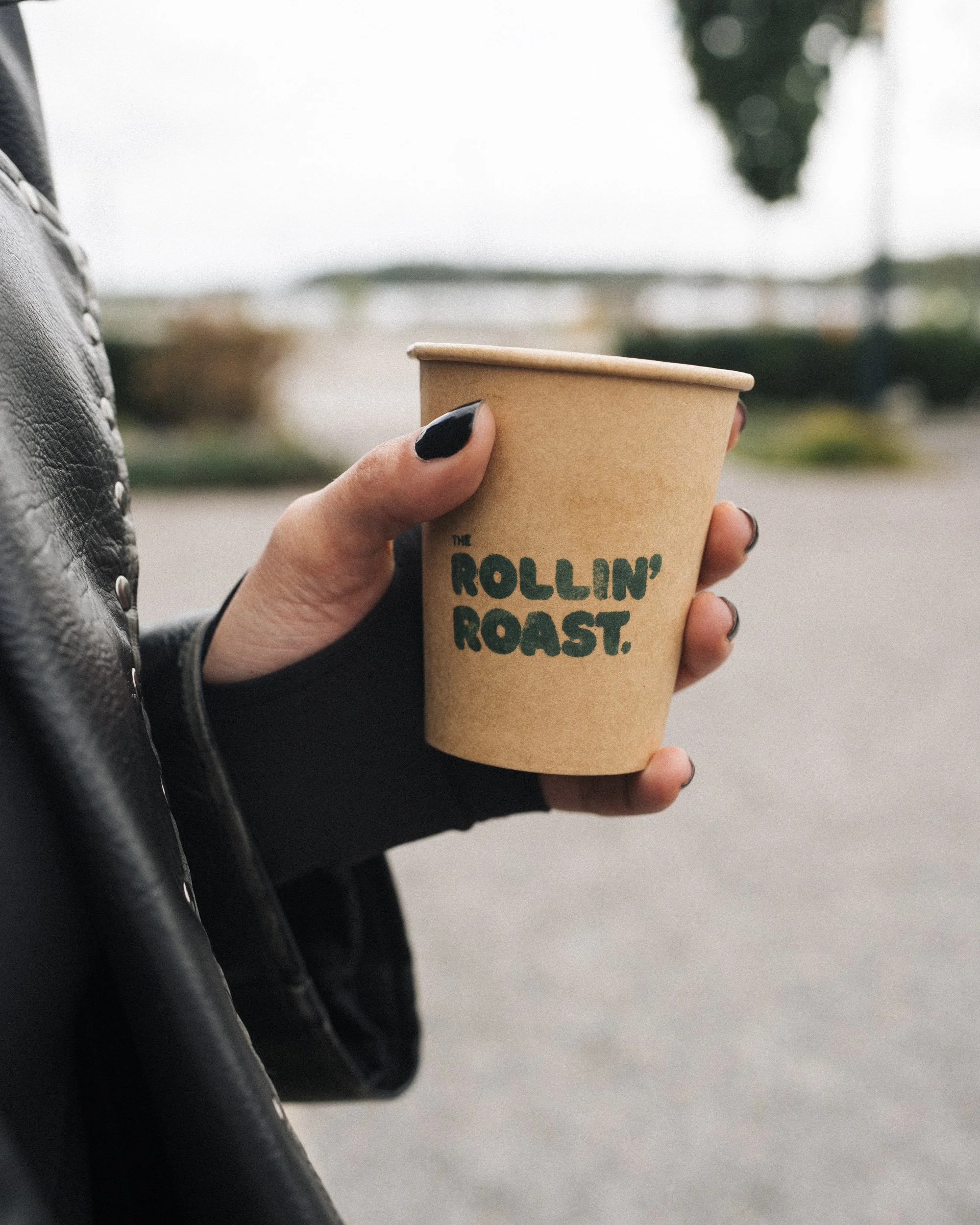 A person holding a disposable coffee cup with the text 'The Rollin' Roost' printed on it, with other guests seated at outdoor tables in the background.