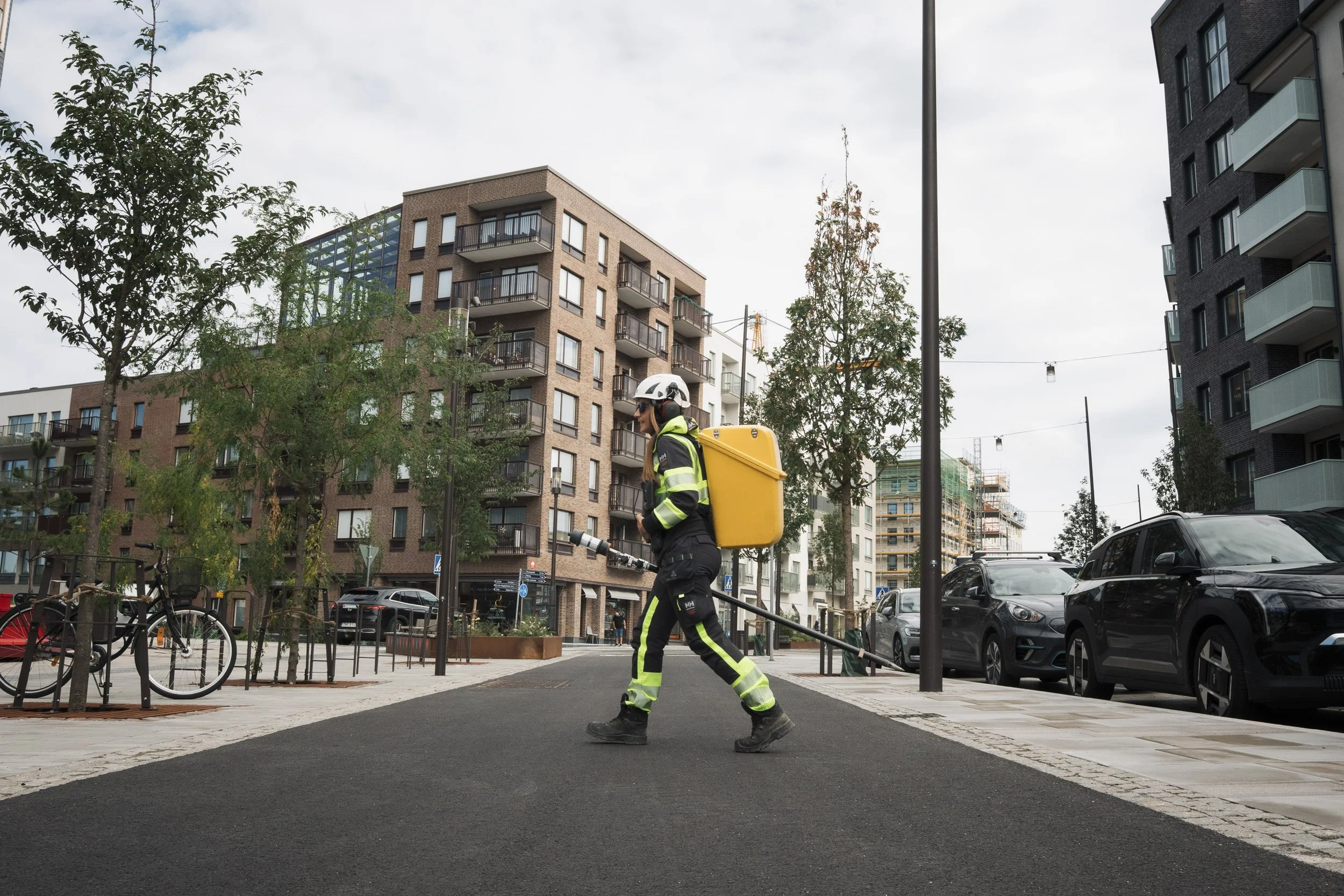 A street scene with a worker in a neon green safety jacket and helmet, carrying a yellow container, walking along the sidewalk, with residential buildings and parked cars in the background.