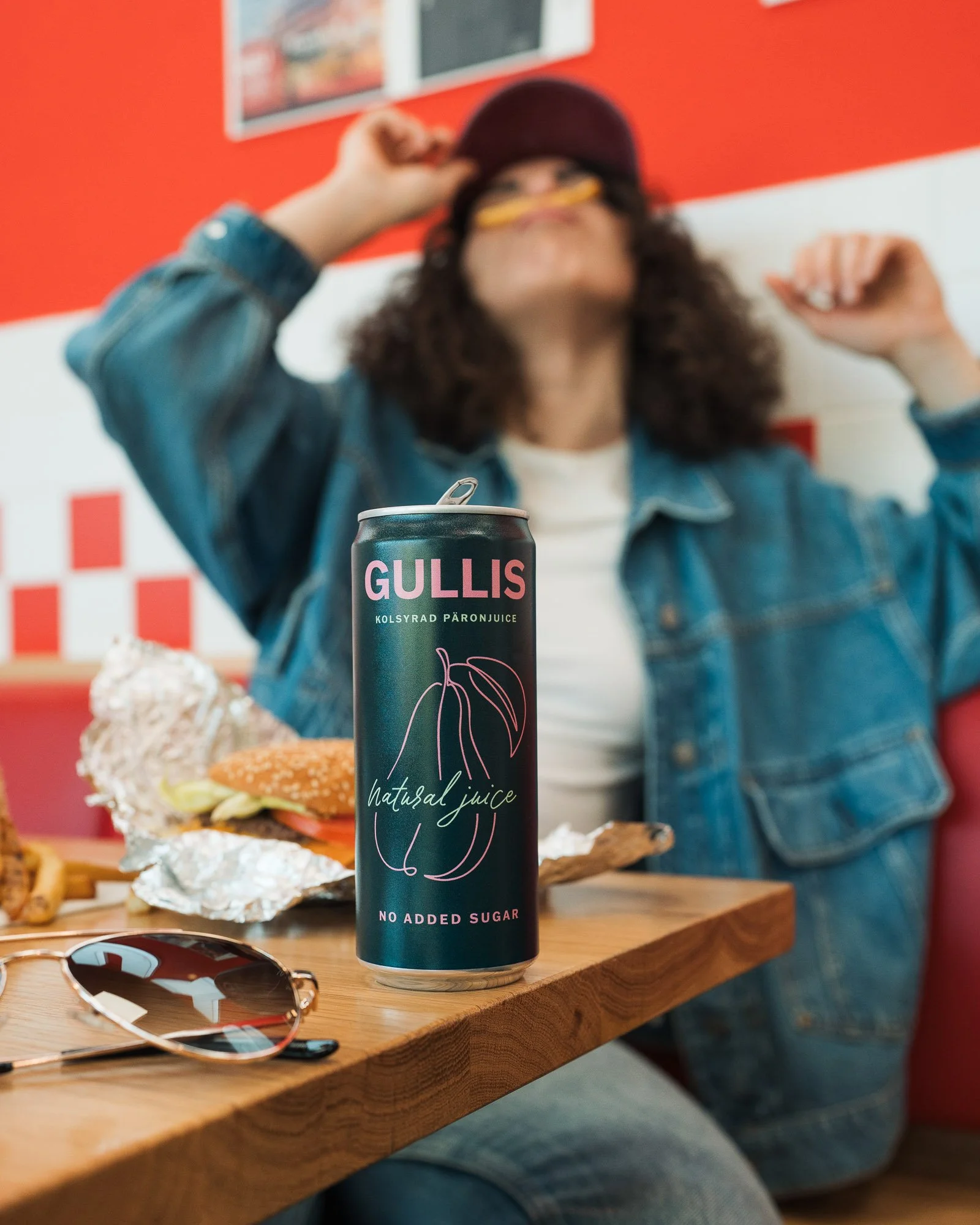 A can of Gullis natural juice with no added sugar placed on a wooden table, with fast food items like a burger and fries in foil wrapping nearby, and a person wearing a denim jacket, white shirt, and red cap seen in the background.