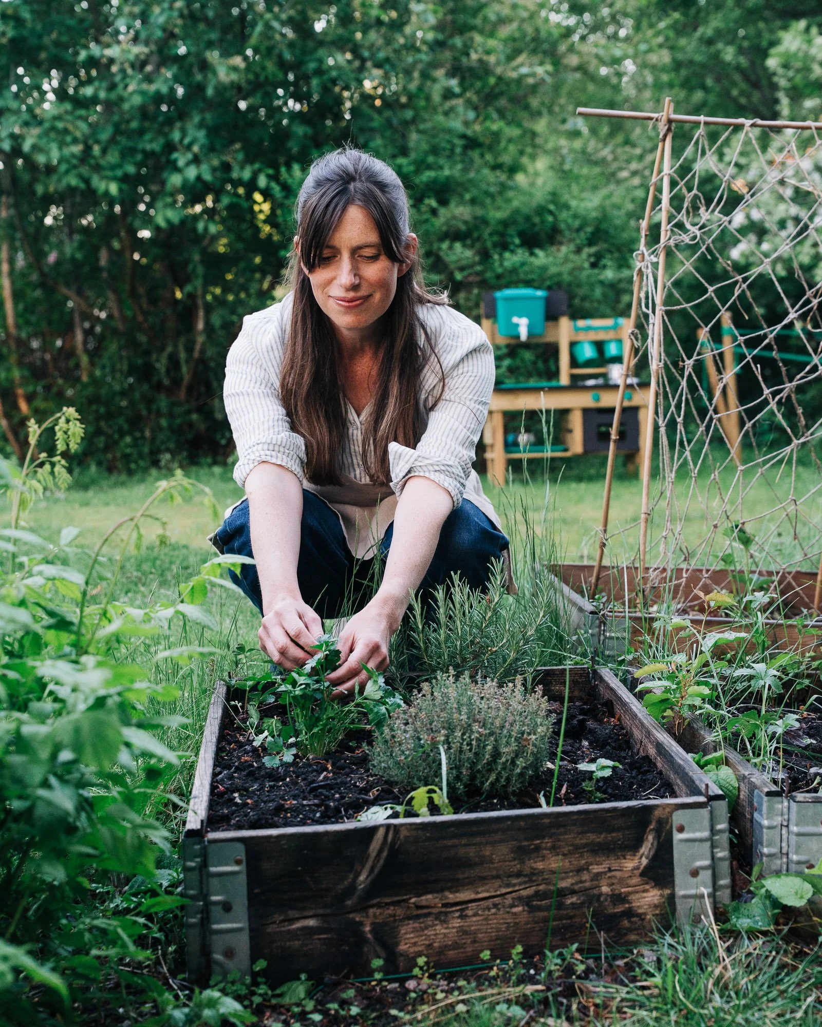 Woman gardening in a backyard, tending to plants in a raised garden bed with a wooden fence and green trees in the background.