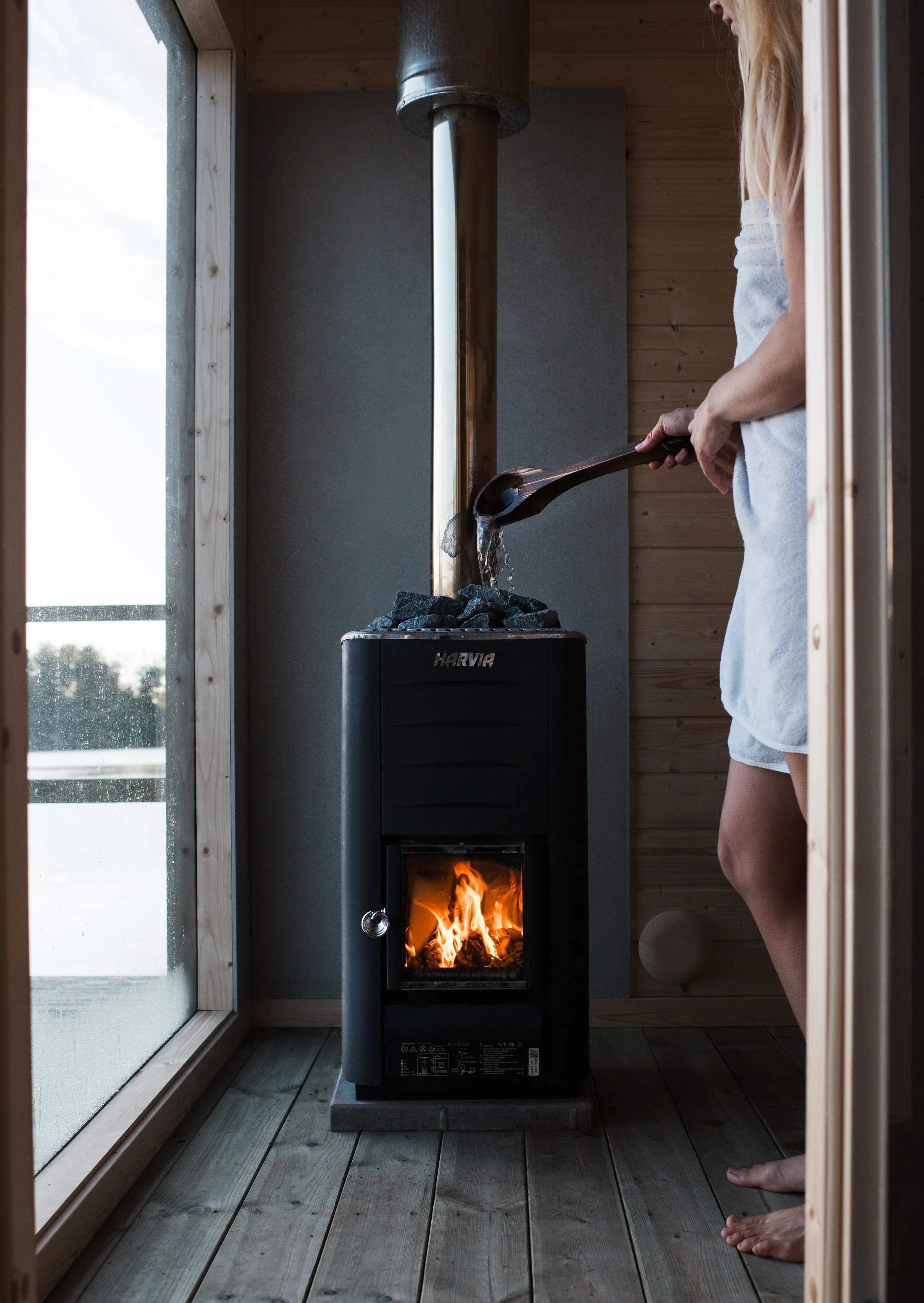 A woman in a white towel standing barefoot on wooden floor, tending a small wood stove with fire and rocks, located in a wooden room next to glass doors showing outdoor natural light.