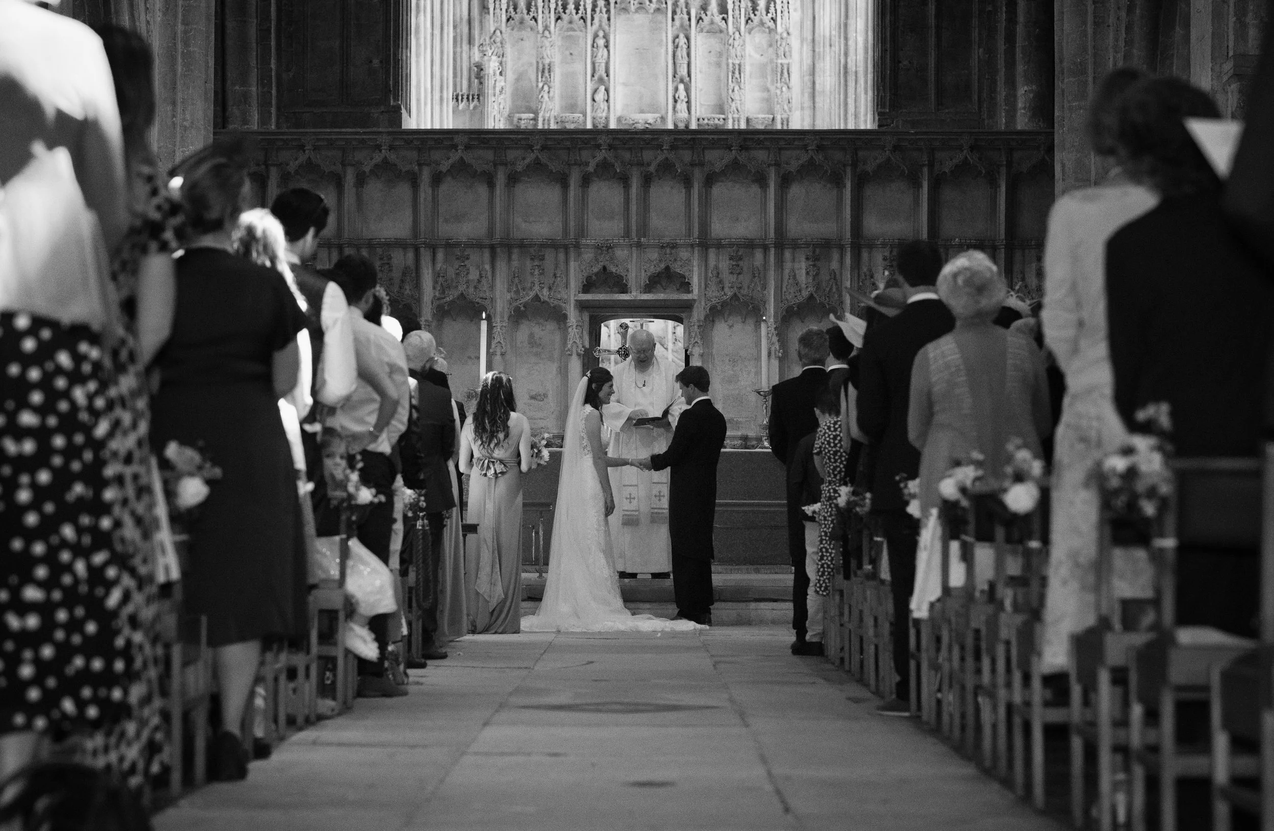 A black and white photograph of a wedding ceremony inside a church, with the bride and groom standing at the altar, surrounded by seated guests.