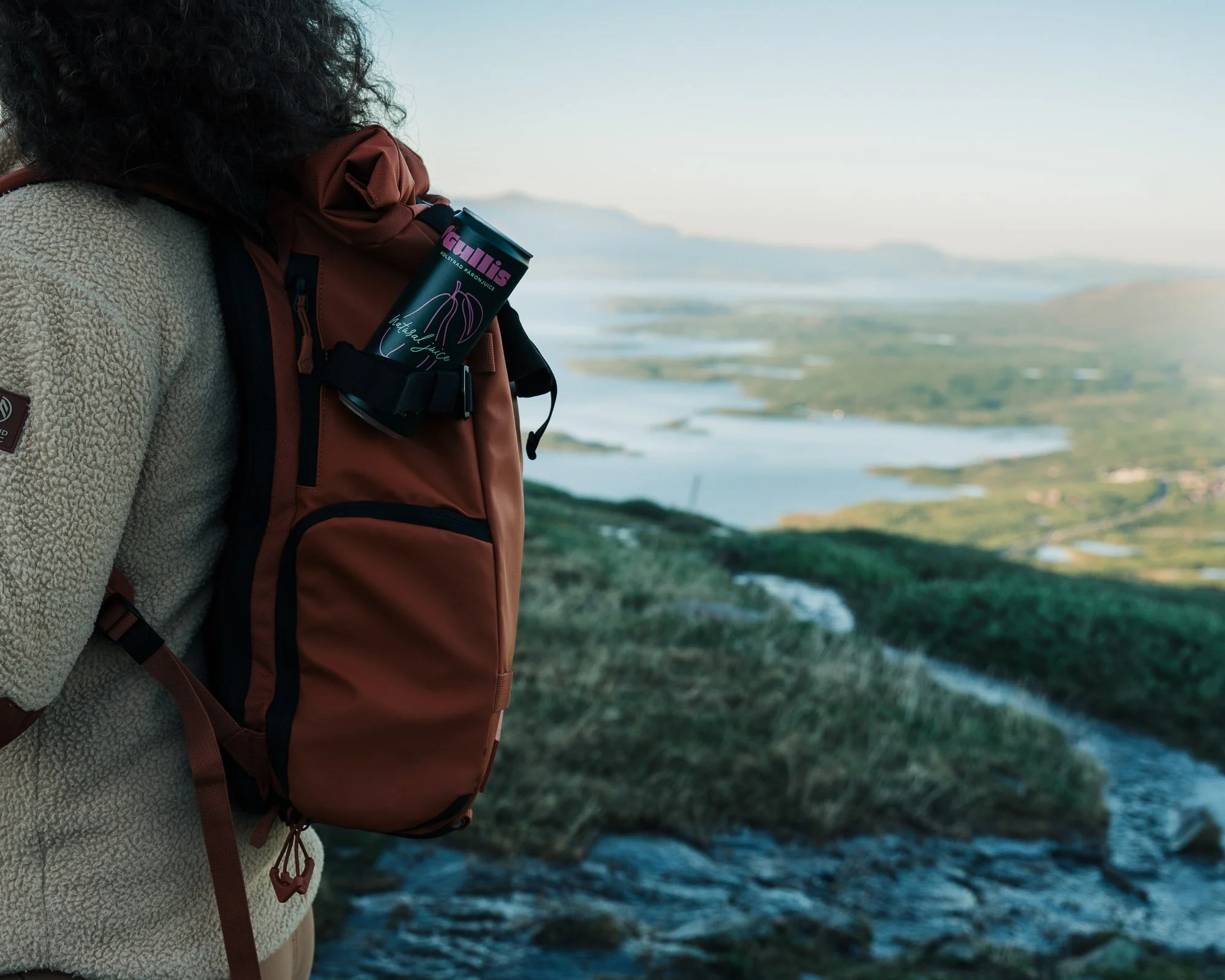Person wearing a beige fleece jacket and a brown hiking backpack, holding a can of juice with text 'Gullis' and a graphic of a melon, overlooking a landscape of water, land, and mountains with a cloudy sky.
