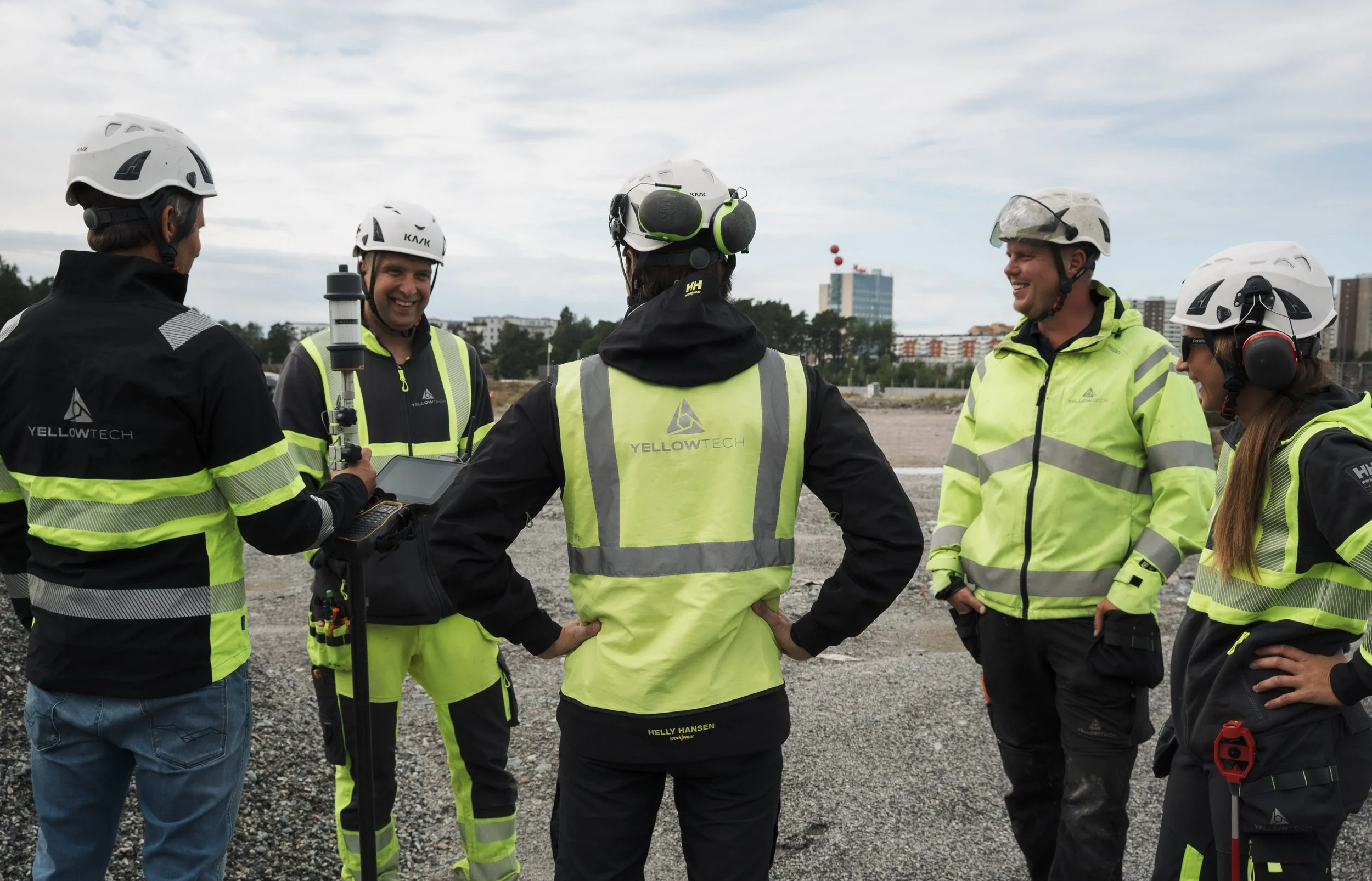 A group of five construction workers wearing safety helmets and high-visibility jackets, standing outdoors on a construction site, engaged in conversation and smiling.