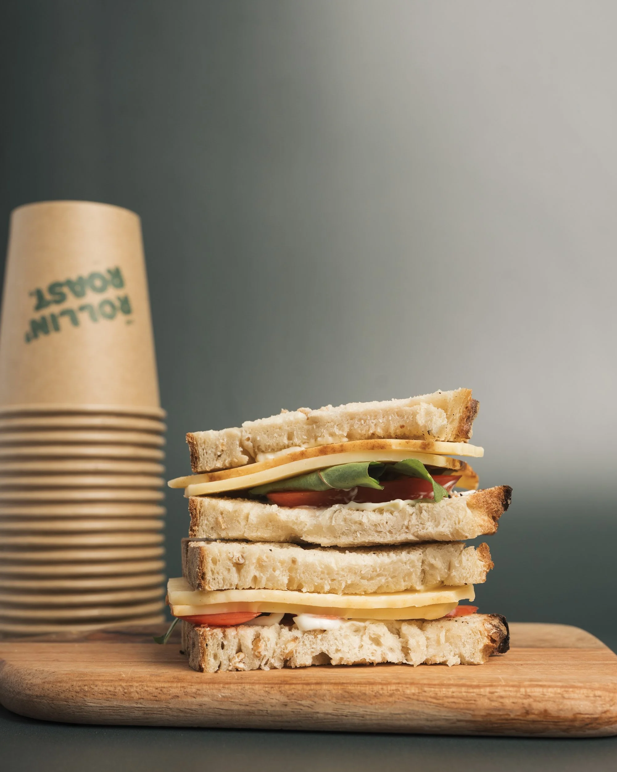 Stacked turkey and cheese sandwiches on a wooden cutting board with lettuce and tomato, with a container of disposable cups labeled 'Rolling Roast' in the background.