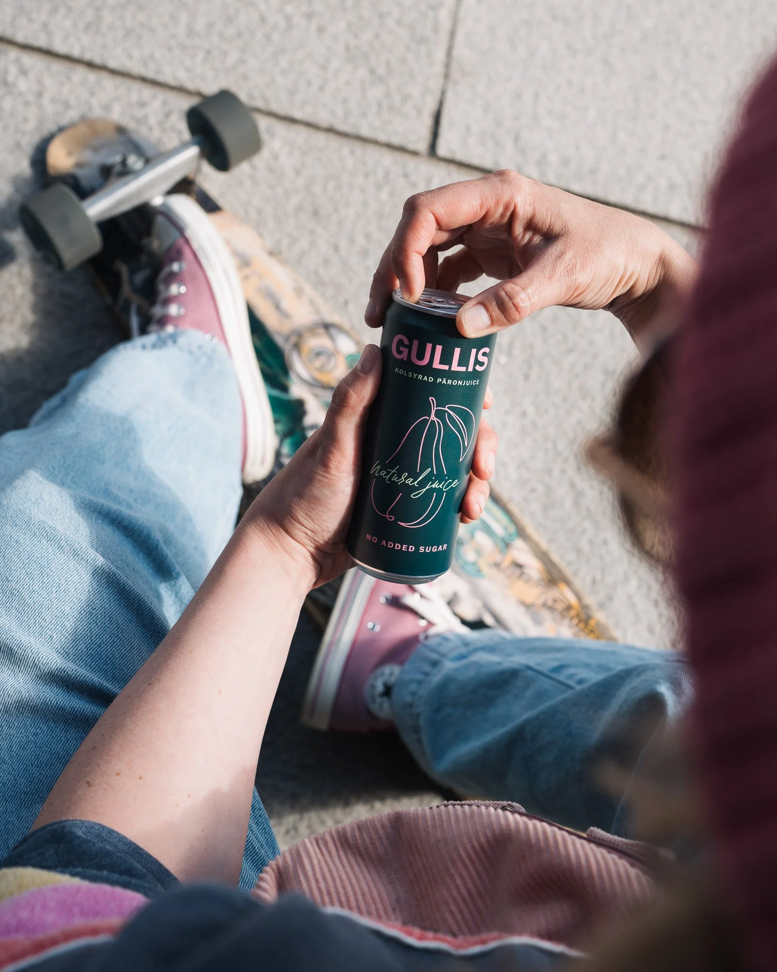 A person sitting on a skateboard with a can of Gullis brand sparkling pomegranate juice, wearing light blue jeans and pink shoes, with another person beside them wearing a pink sweater. The photo is taken outdoors on a concrete surface.